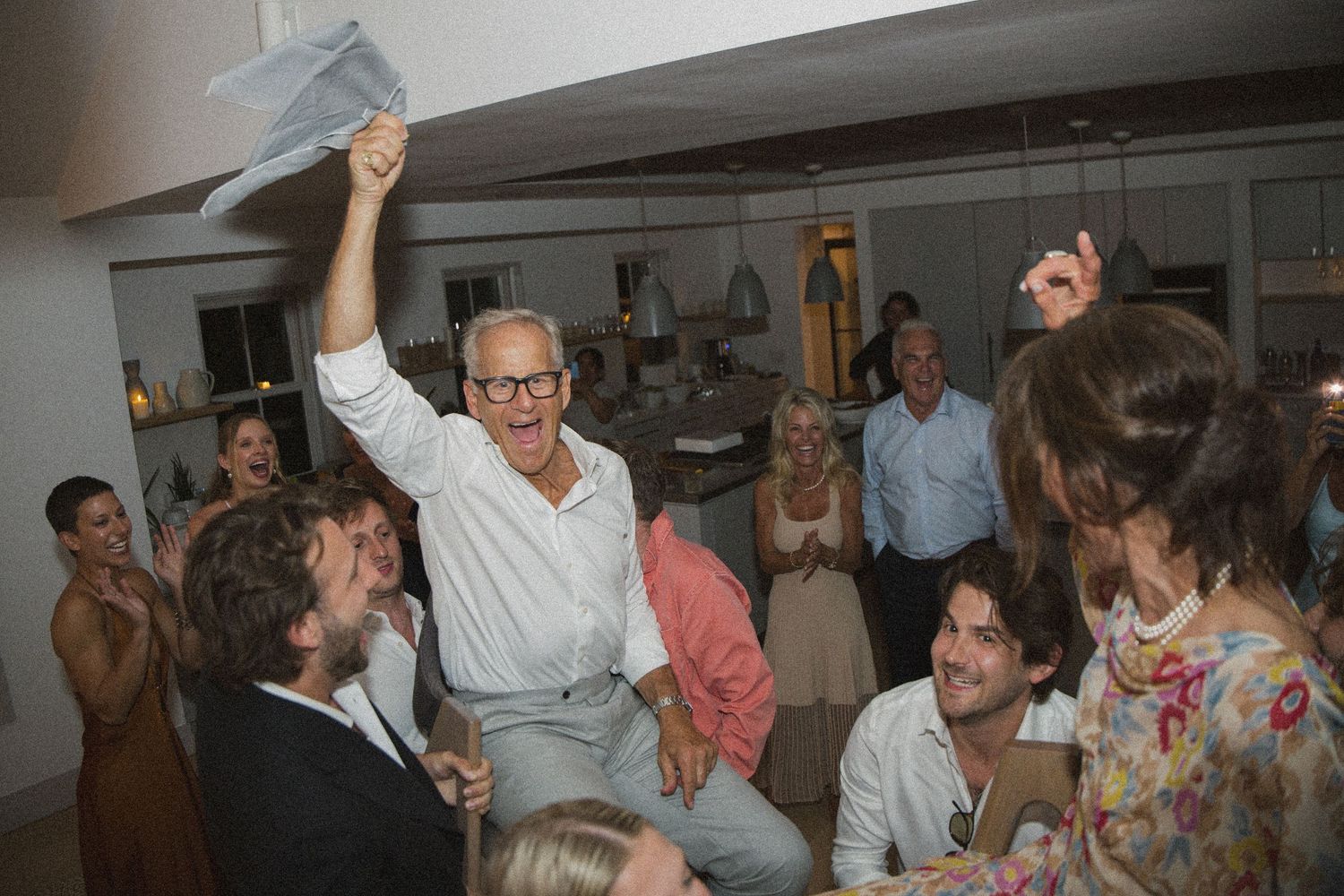 Elderly man joyfully raises paper at lively indoor party surrounded by smiling guests in casual attire.