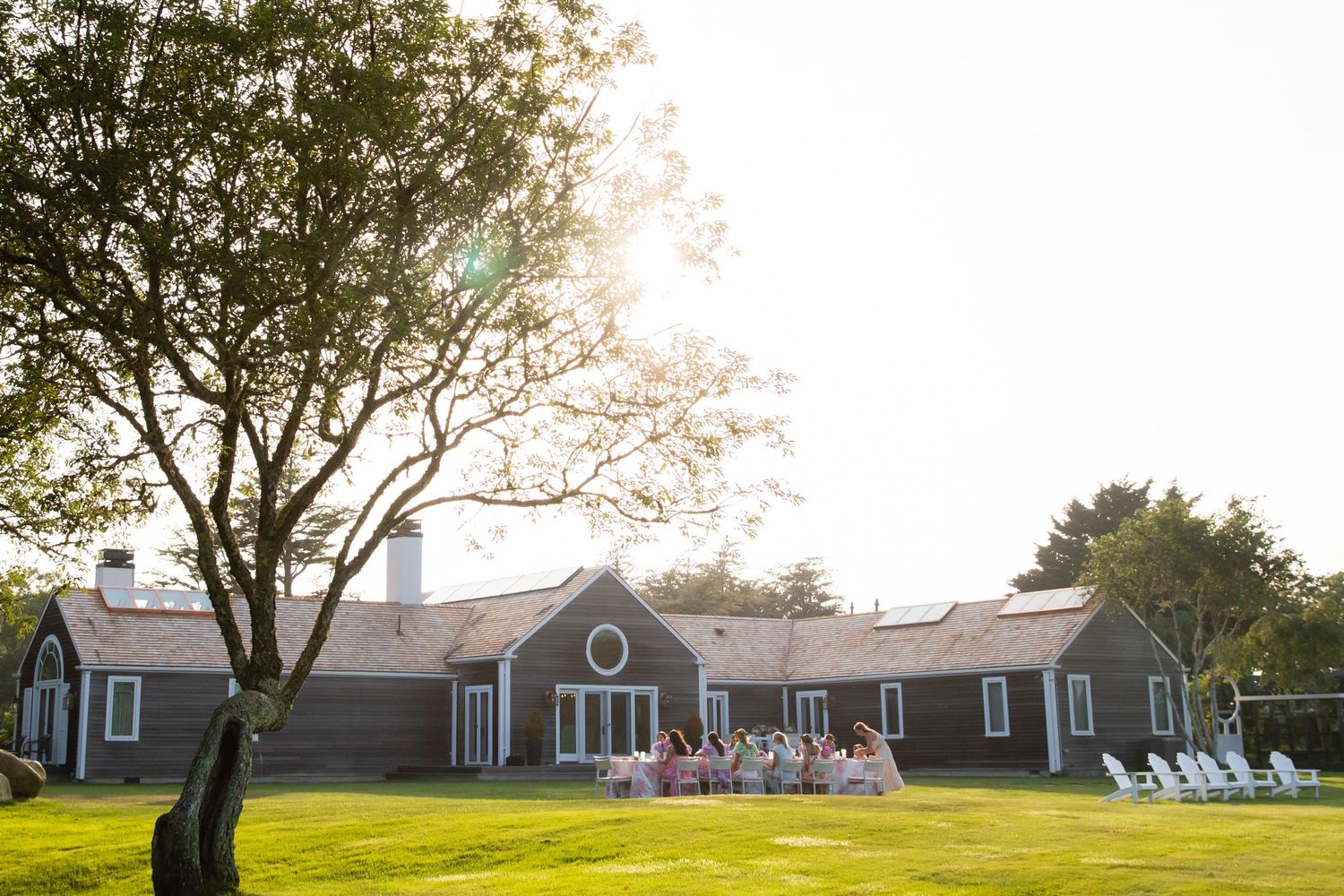 Outdoor wedding reception on lush green lawn beside charming gray New England barn venue at golden hour sunset.