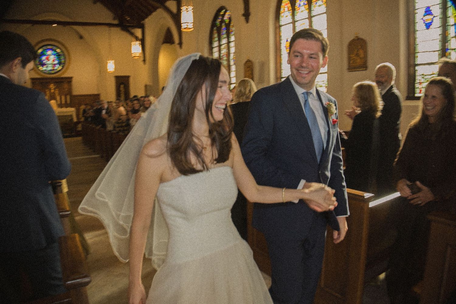 Smiling bride in a white gown and veil holds hands with her groom in a navy suit as they walk down the church aisle past wedding guests.