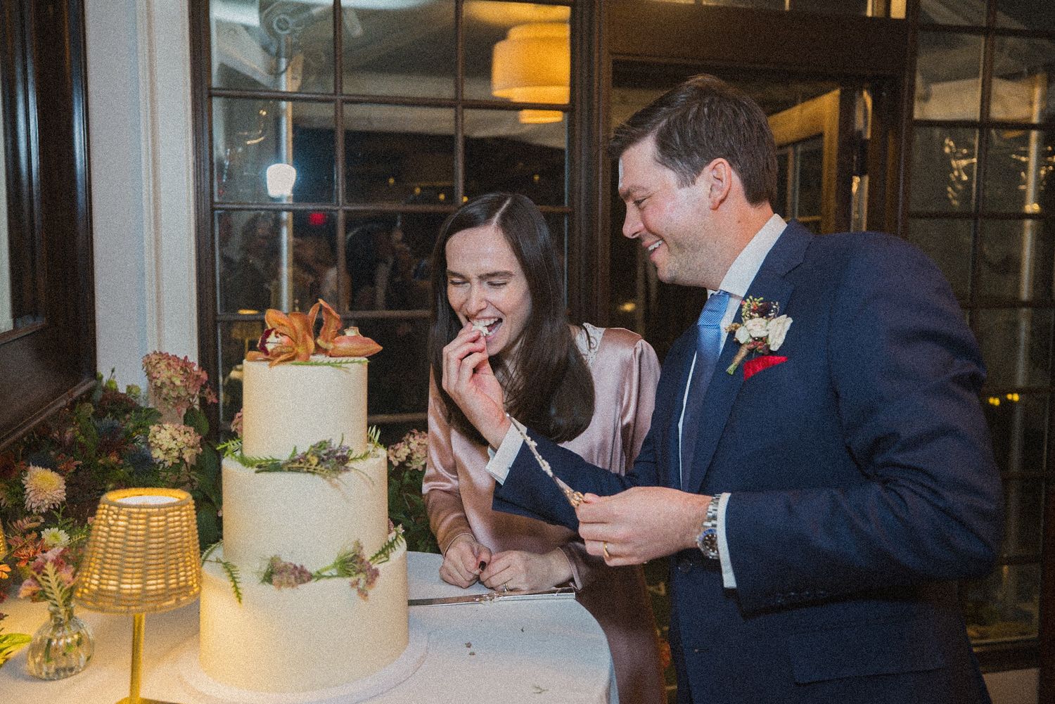 A smiling couple tastes a three-tier white wedding cake decorated with greenery and flowers at an elegant indoor reception.