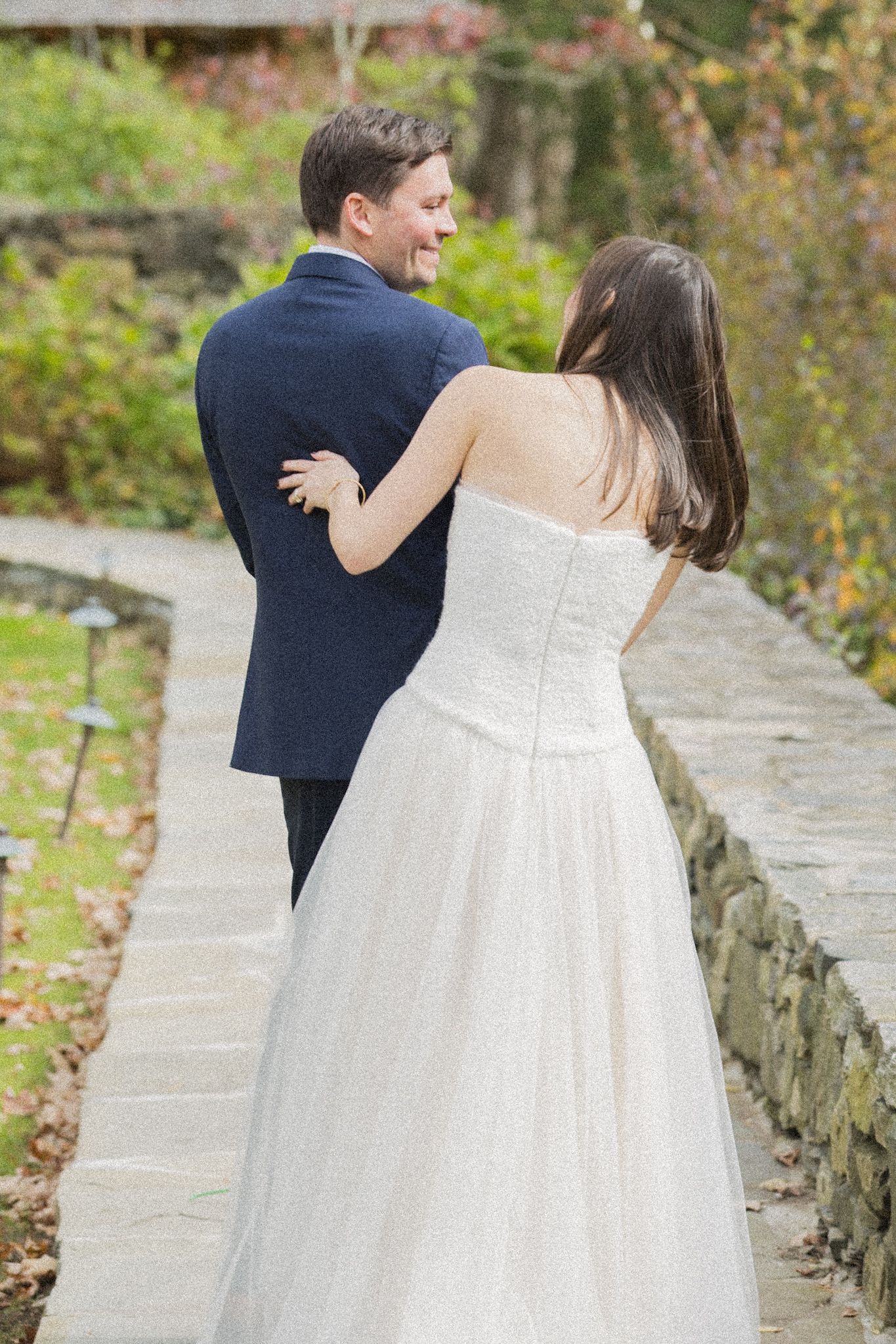 Bride in a strapless white tulle gown rests her hand on a smiling groom in a navy suit as they walk a stone garden path in autumn.