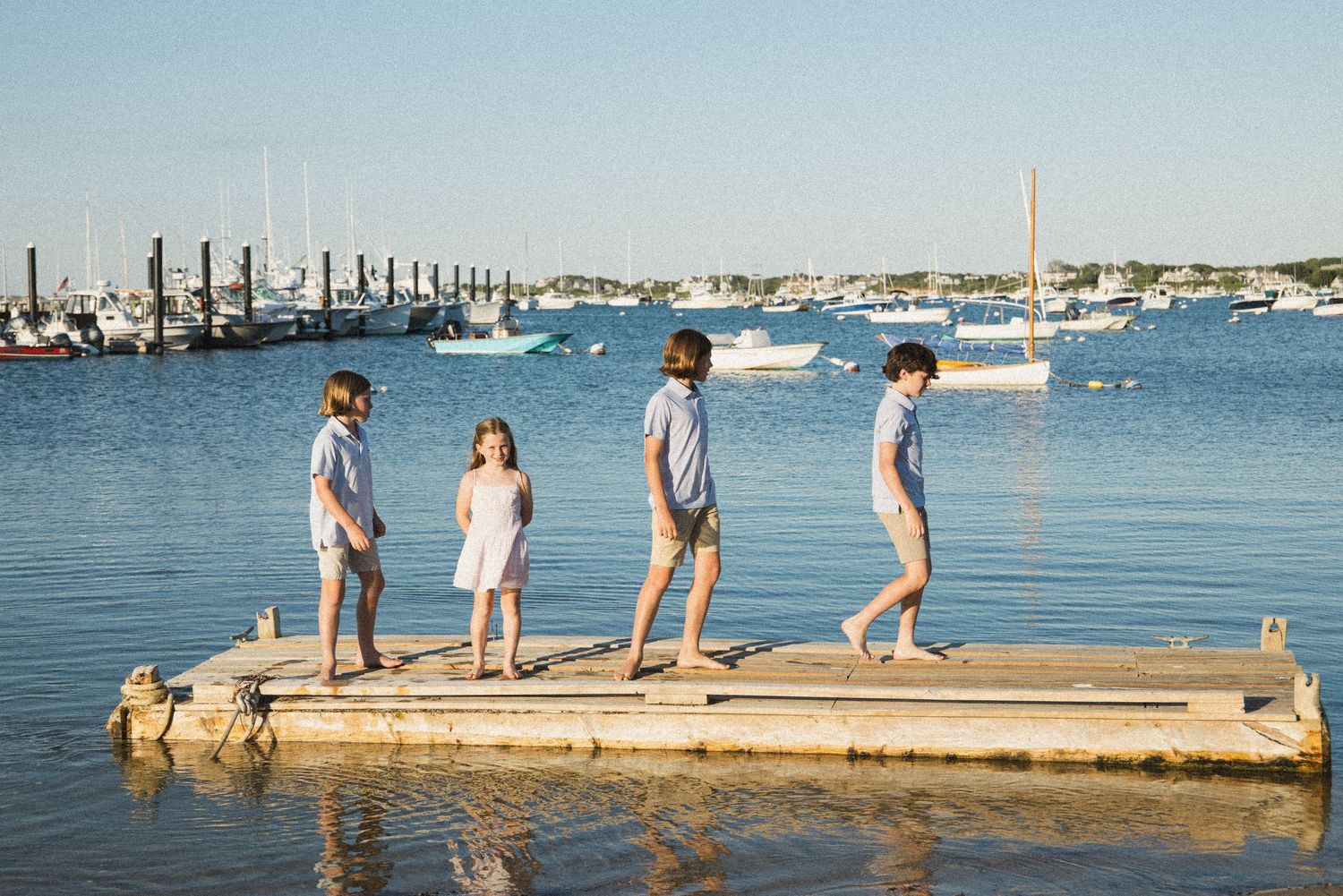 Four children walking on a wooden dock with sailboats and calm harbor waters in the background on a sunny day.