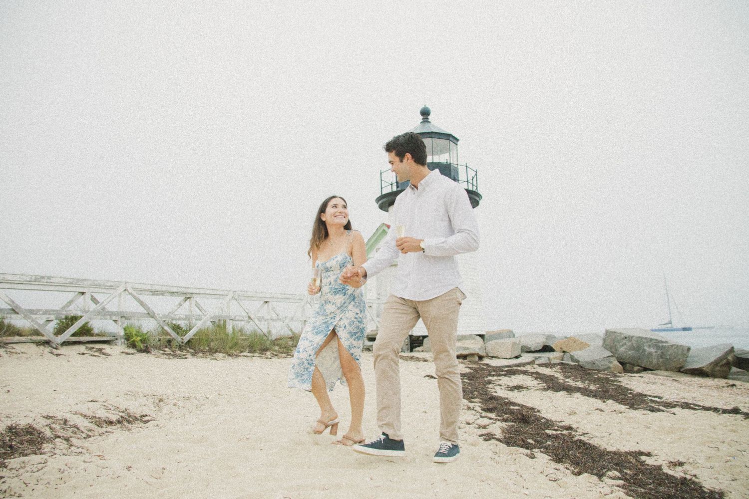 Couple walking hand in hand on a sandy beach near a lighthouse on a foggy day.