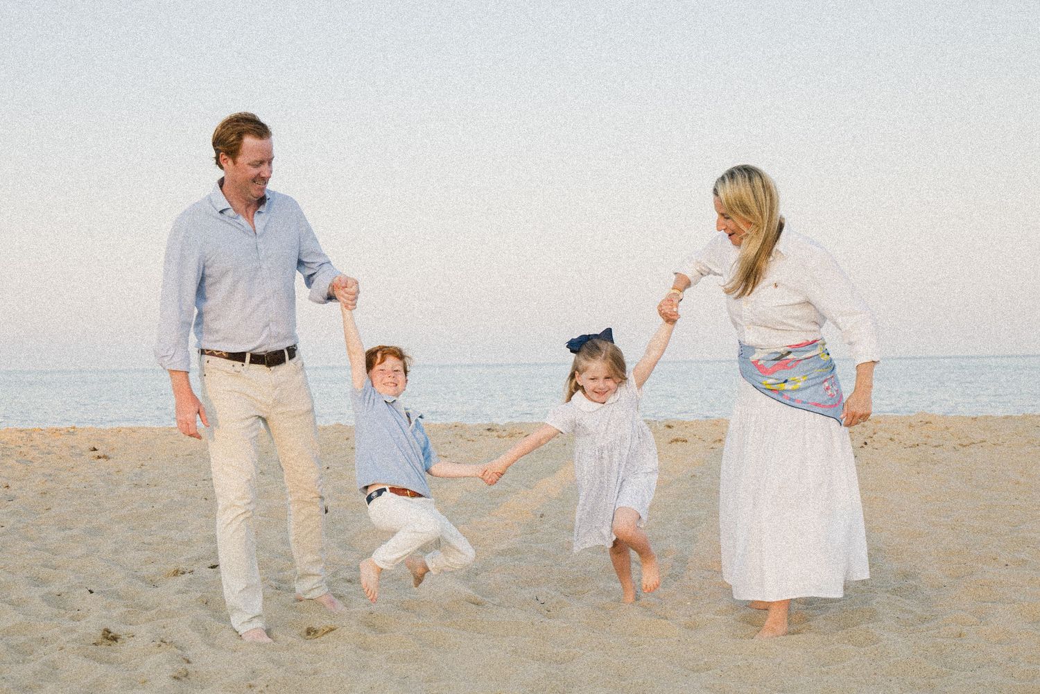 Family of four playing on a sandy beach, parents swinging two young children by their hands near the ocean shoreline.