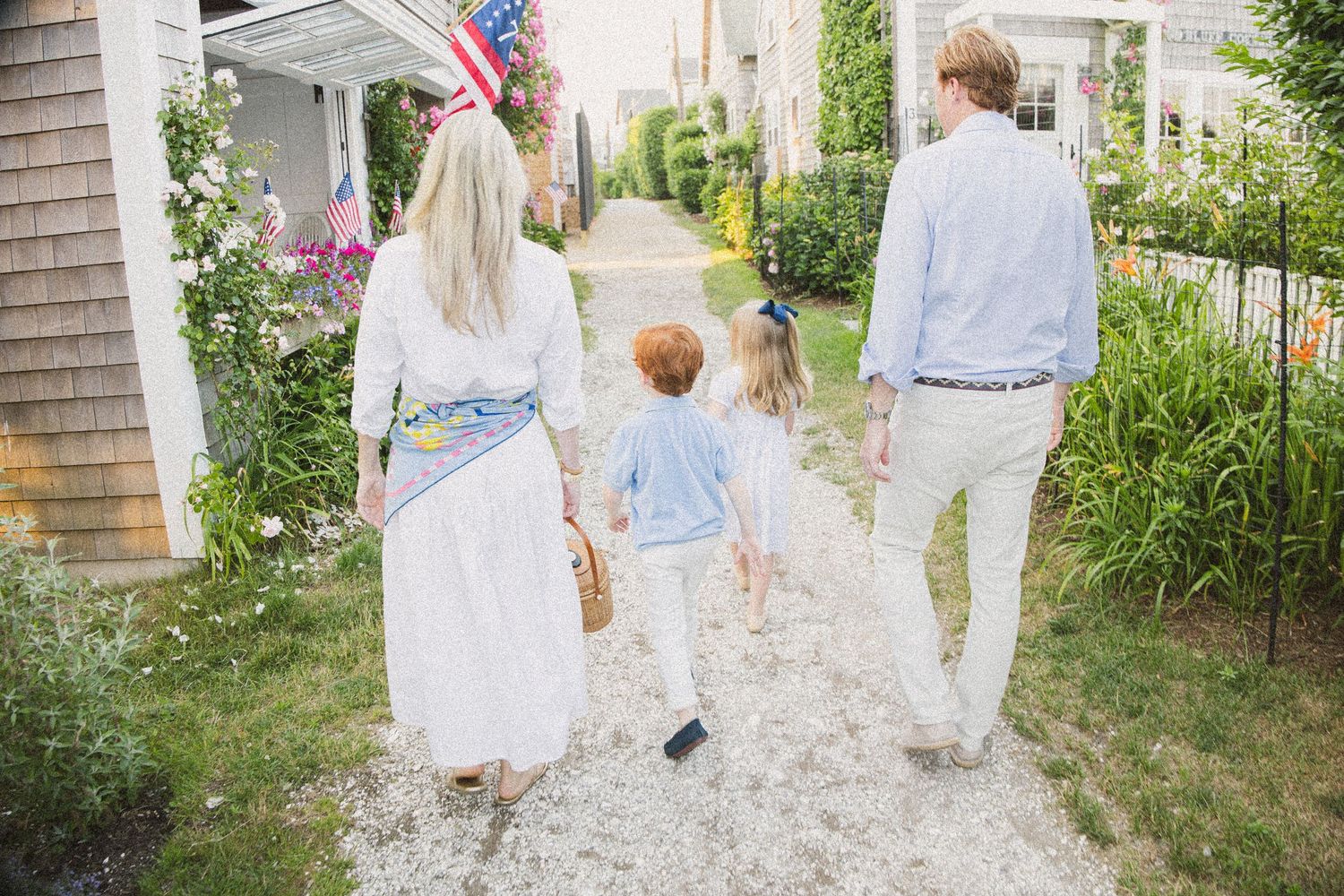 Family of four walking hand-in-hand down a charming garden path past an American flag-adorned home.
