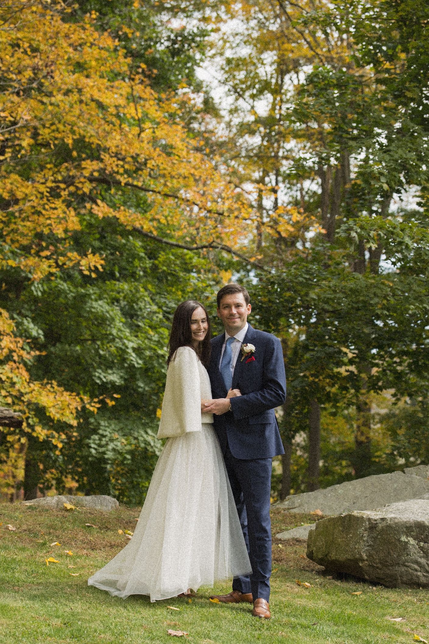 Smiling bride in a white tulle skirt and shawl embraces a groom in a navy suit on a lawn surrounded by autumn trees and boulders.