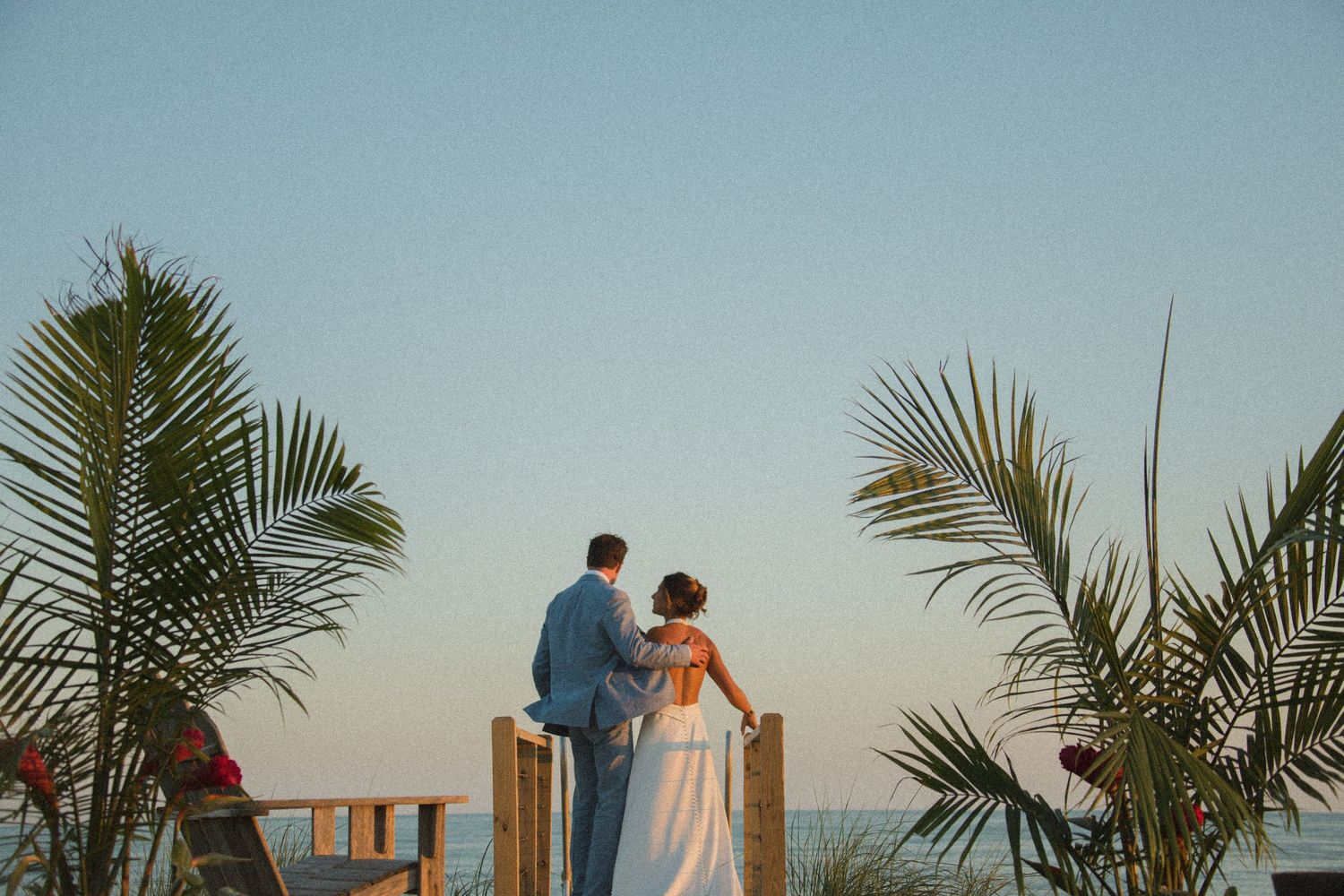 A couple poses near wooden posts and palm trees along a beachfront boardwalk at sunset.