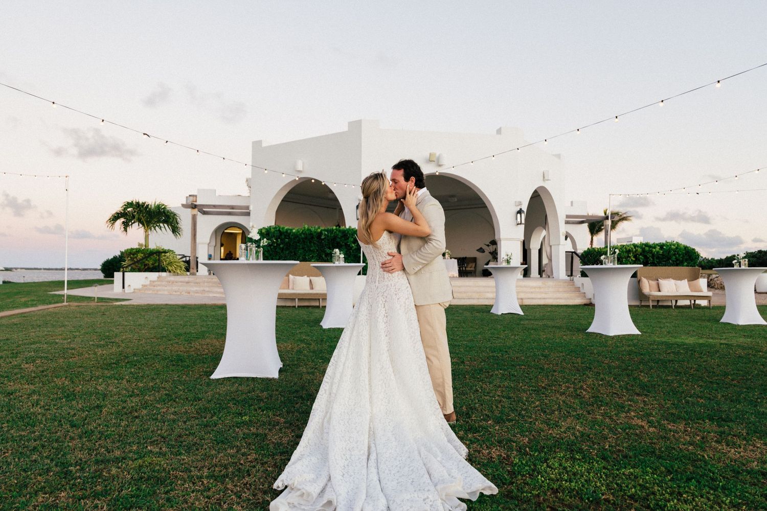 Wedding reception setup on grassy lawn with white decor and ocean views.