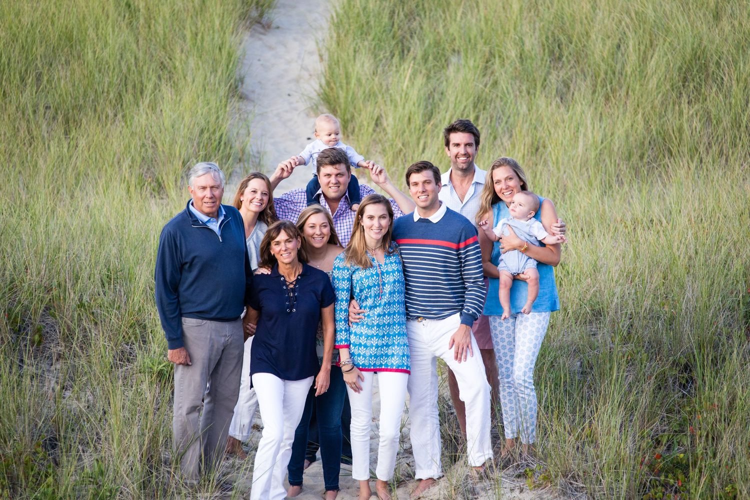 Large family group smiling together on a sandy beach path surrounded by tall green grass on a sunny day.