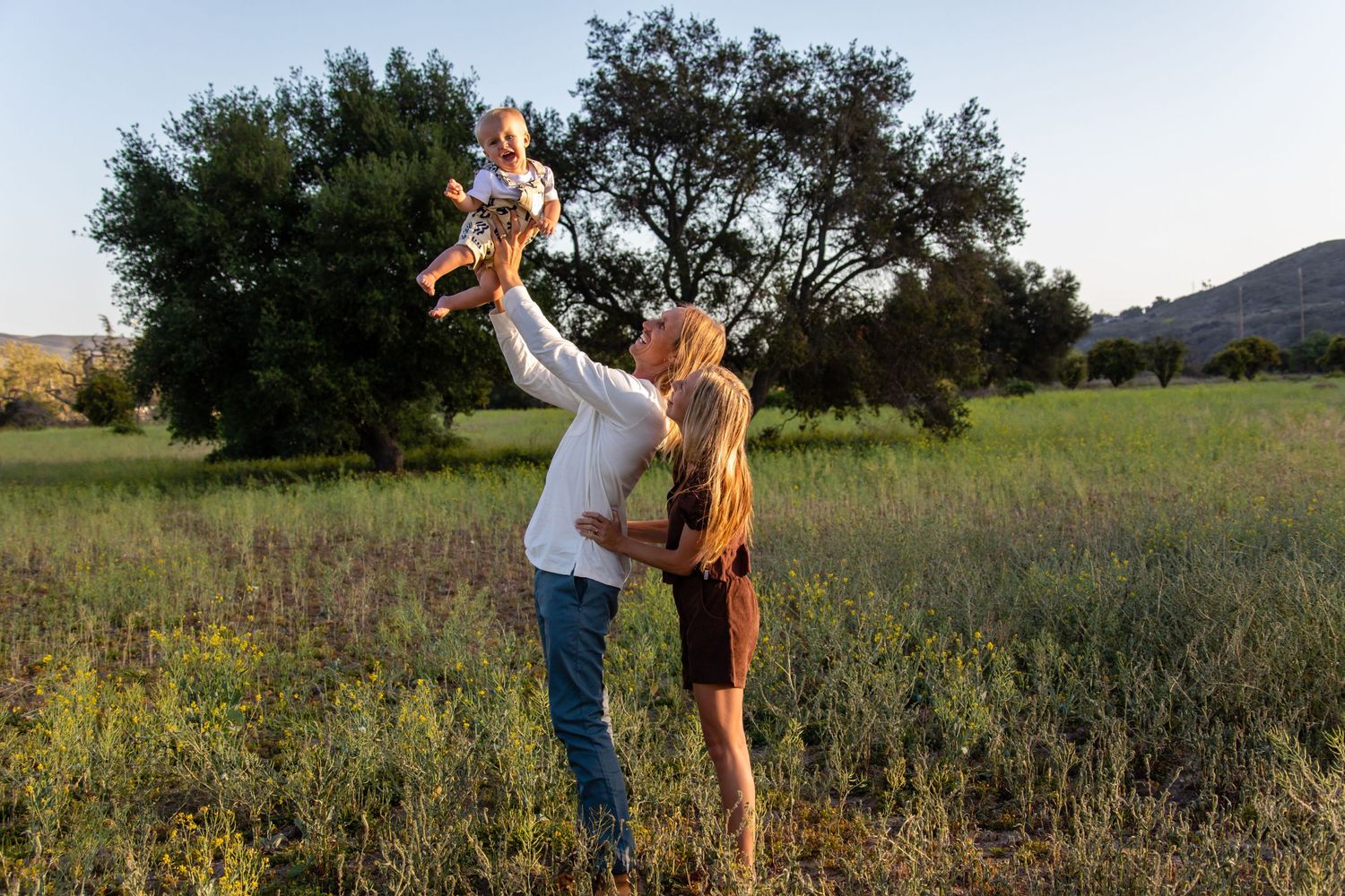 A couple lifts their child in the air while playing in an open field.