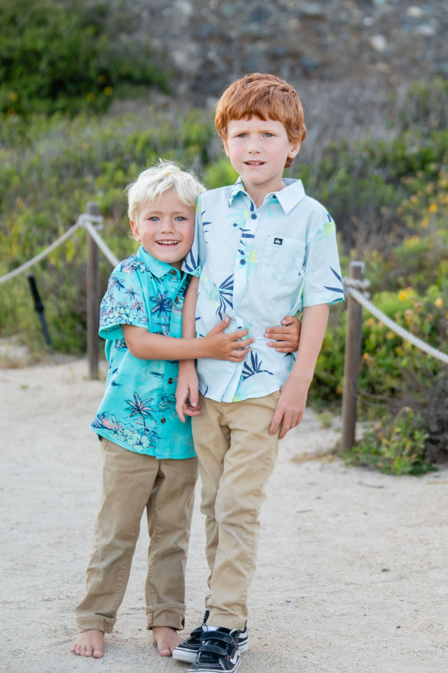 Two young children in tropical print shirts stand together on a dirt path.