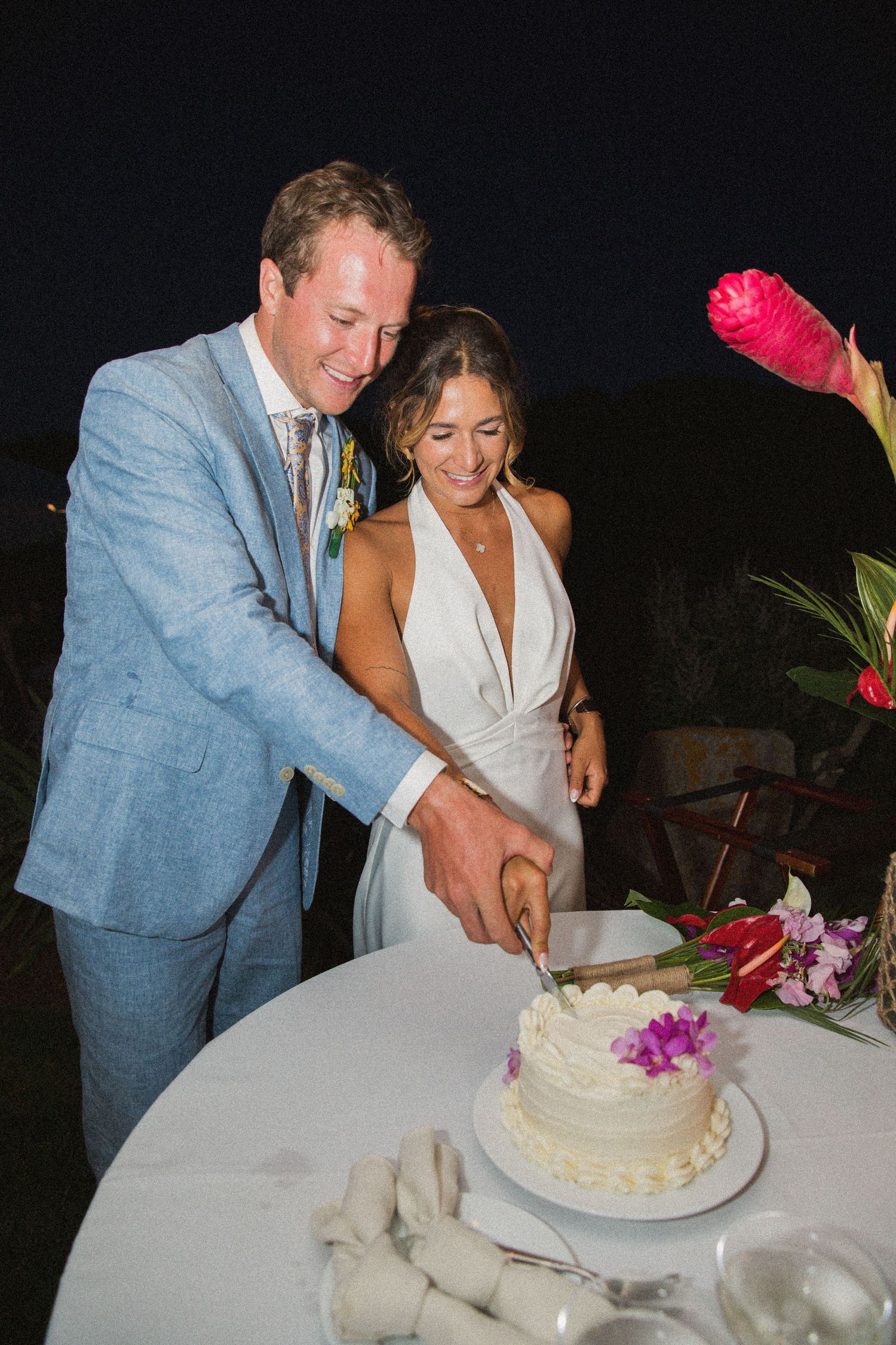 Couple cutting their white wedding cake decorated with purple orchids at an outdoor reception.