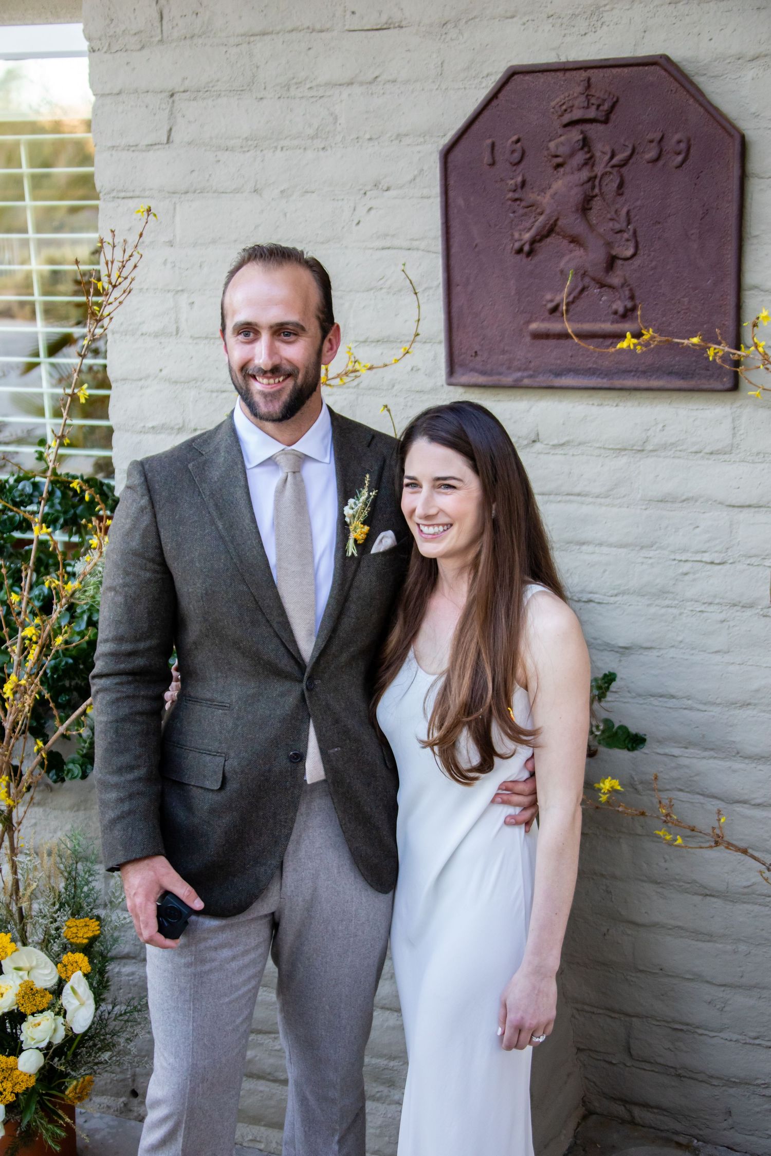 A couple poses against a textured wall with decorative plaques during a wedding photoshoot.