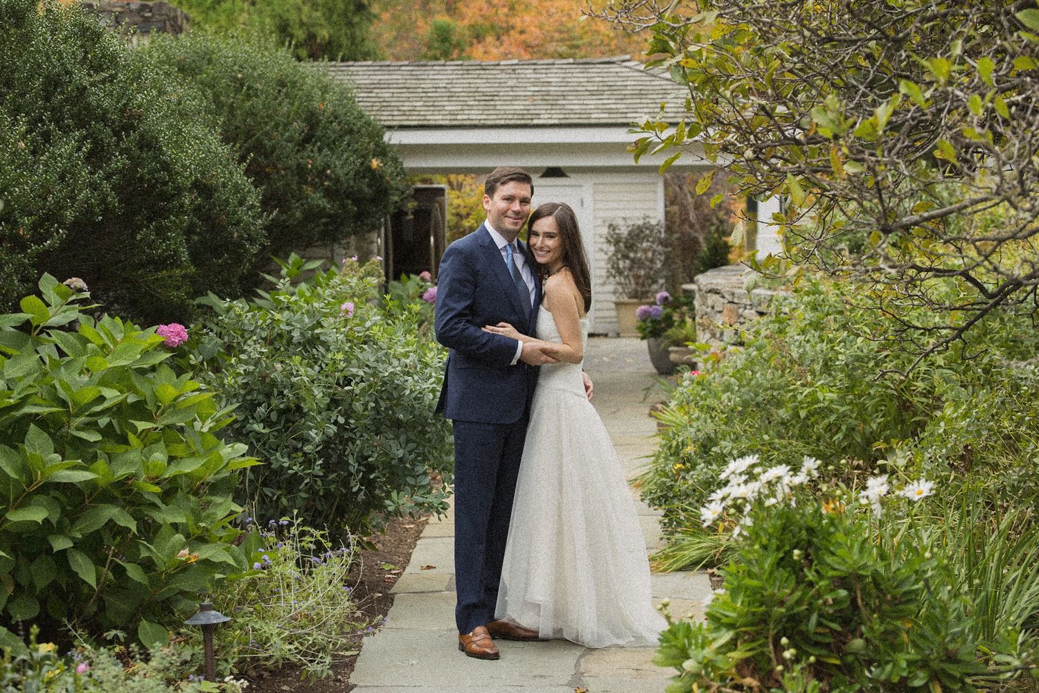 A smiling bride and groom pose together on a garden path surrounded by lush greenery and flowering shrubs in autumn.