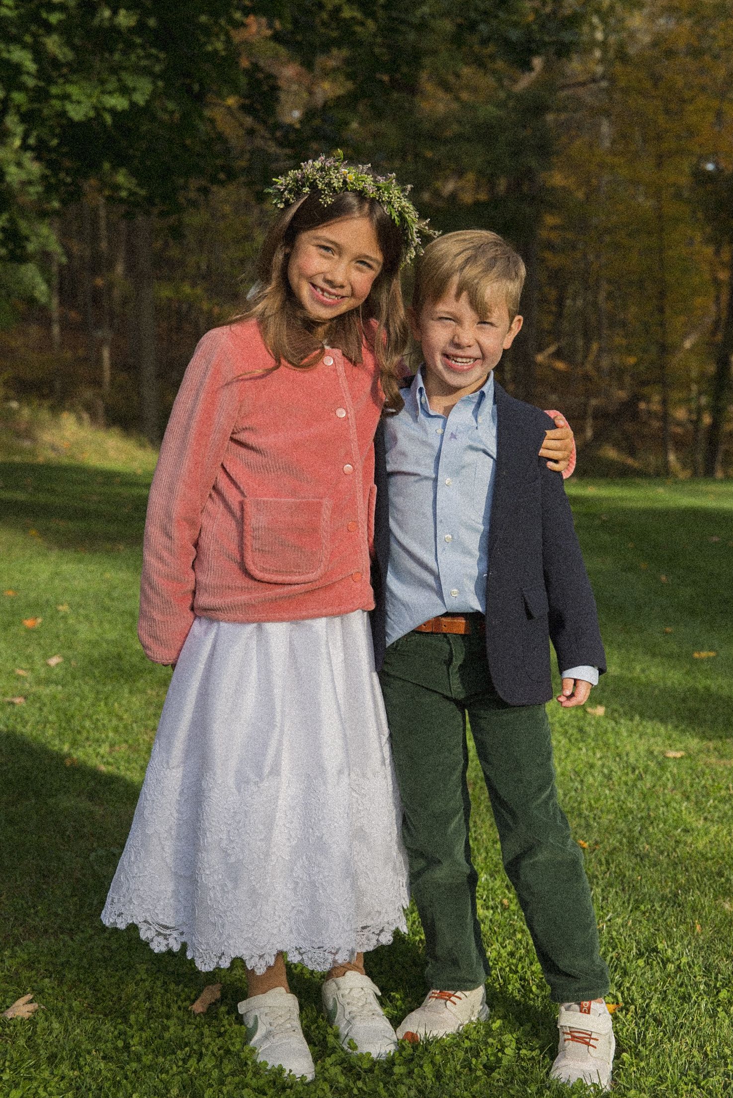Smiling girl in a flower crown and white lace skirt stands beside a laughing boy in a navy blazer and green pants on a sunny lawn.