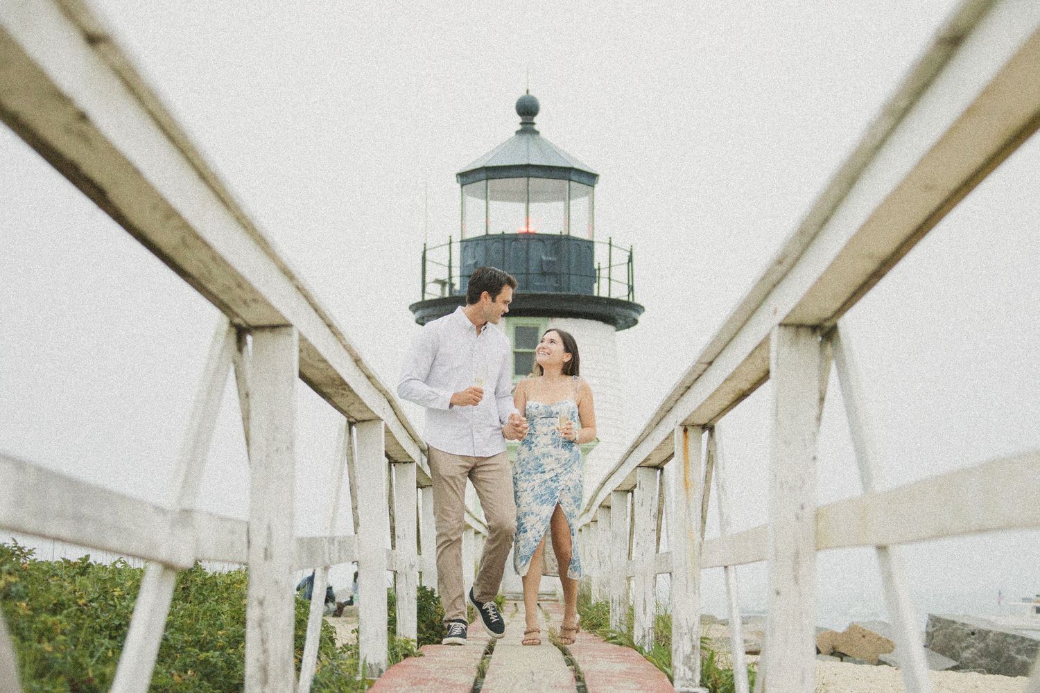 Couple walking hand in hand on a wooden bridge pier with a lighthouse in the background.