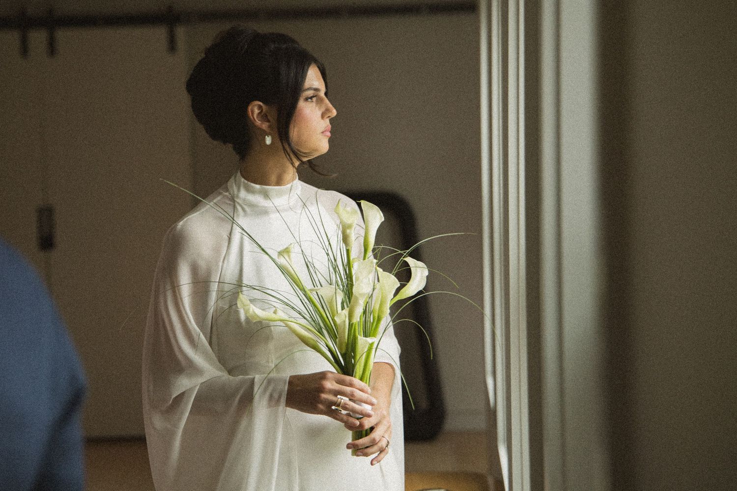 Bride in white dress holding calla lily bouquet gazes out window in elegant bridal portrait.