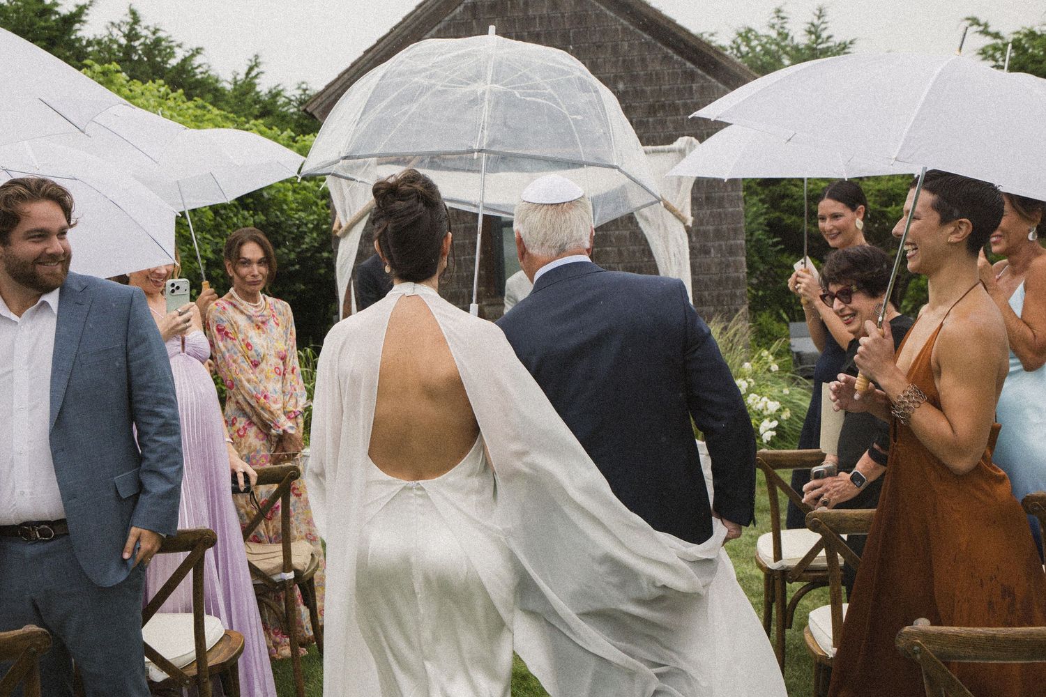 Bride in backless gown walks down outdoor aisle with escort holding clear umbrella at rainy garden wedding ceremony.
