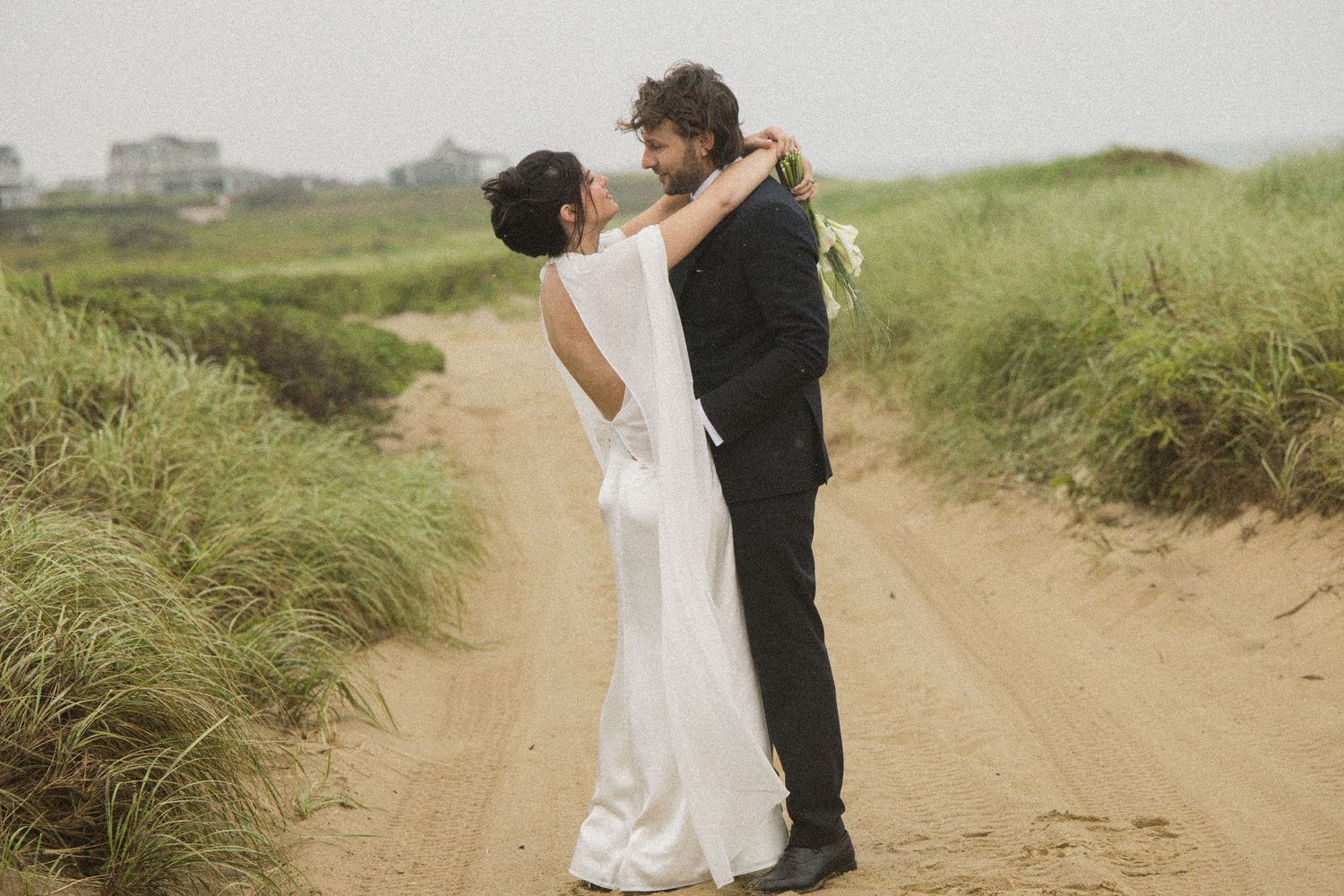 Bride and groom embracing on a sandy coastal path surrounded by green dunes on their wedding day.
