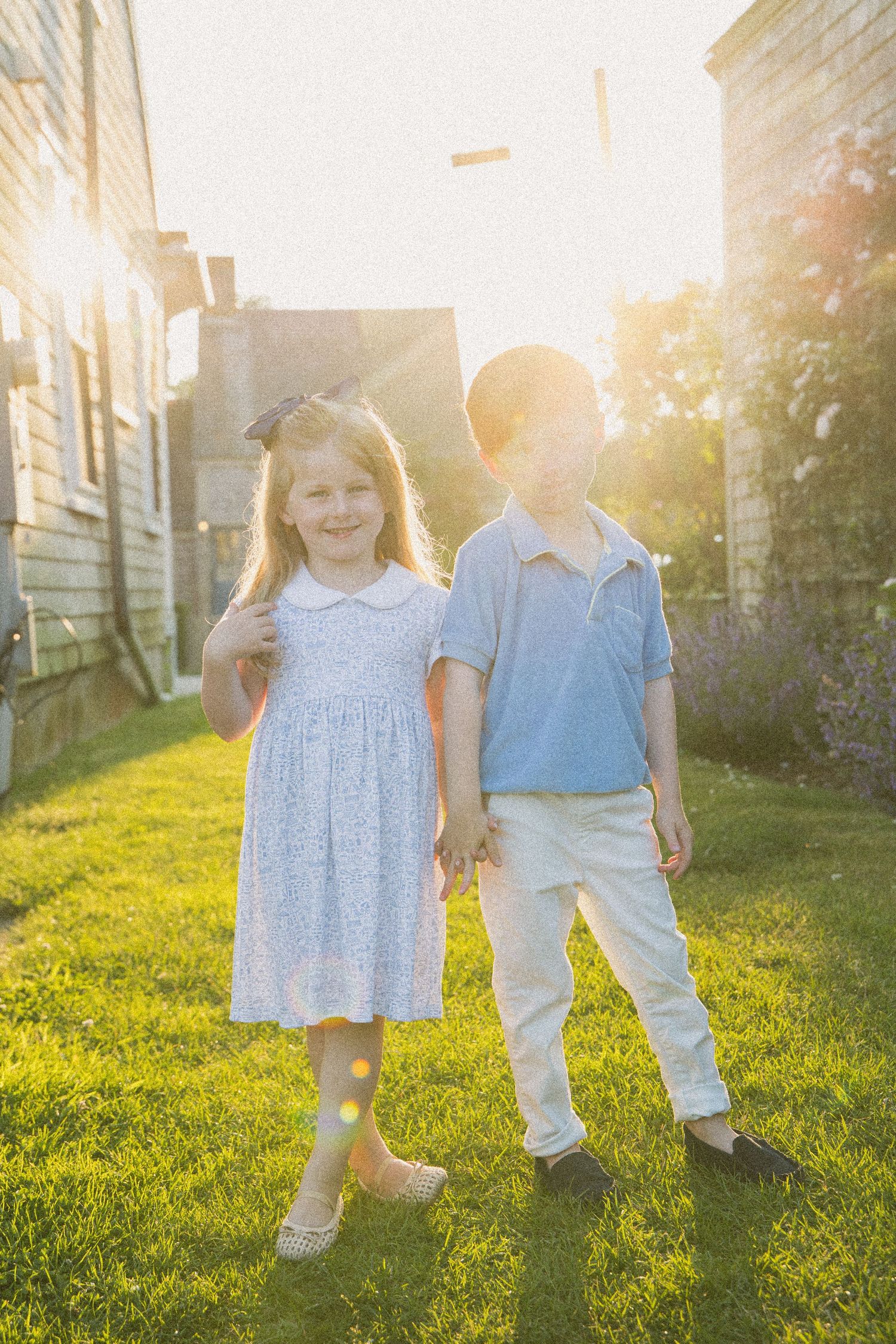 Two young children holding hands on green grass in golden sunset light, girl in white dress, boy in blue polo shirt.