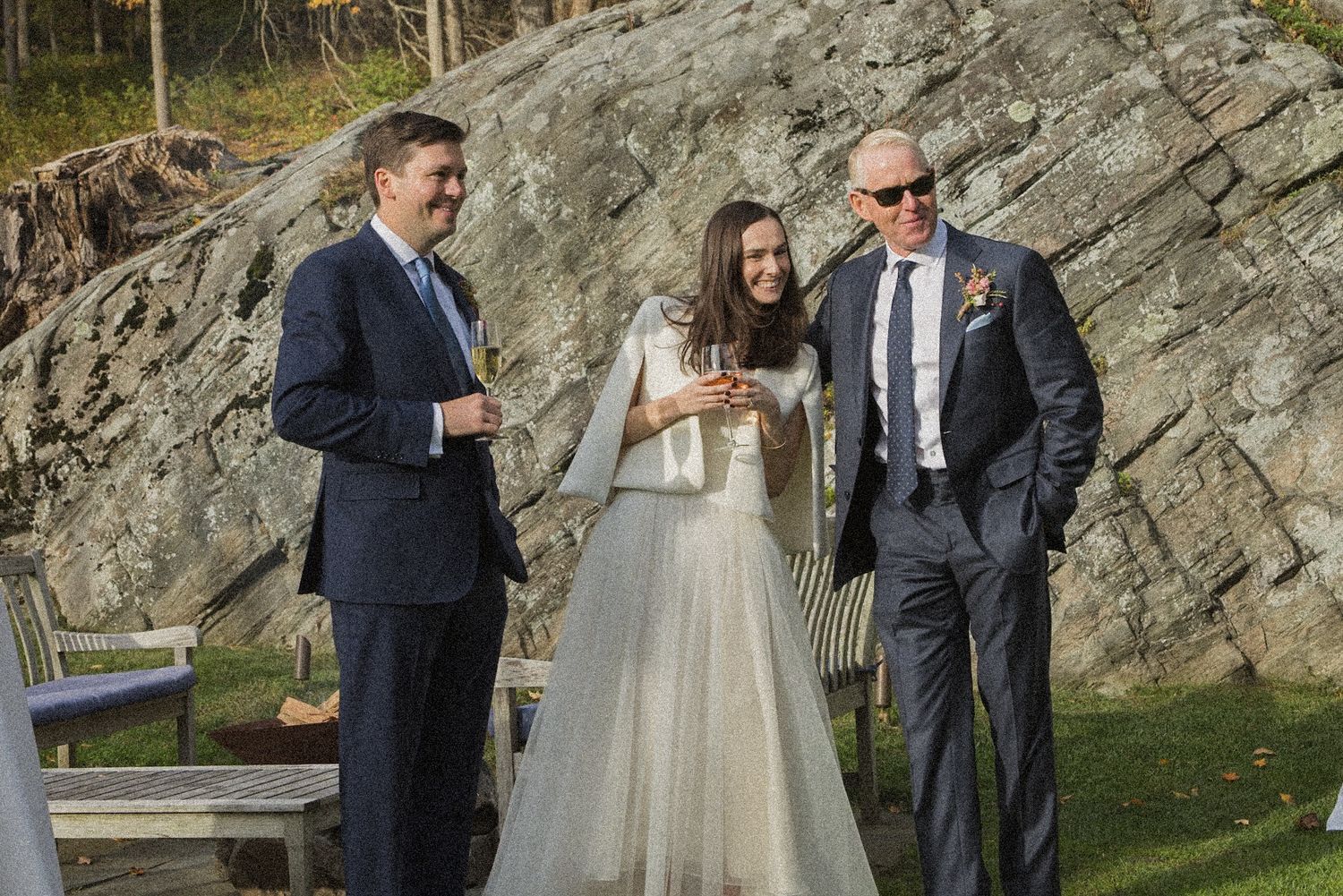 A bride in a flowing white gown stands outdoors between two men in dark suits in front of a large rock face on a sunny day.