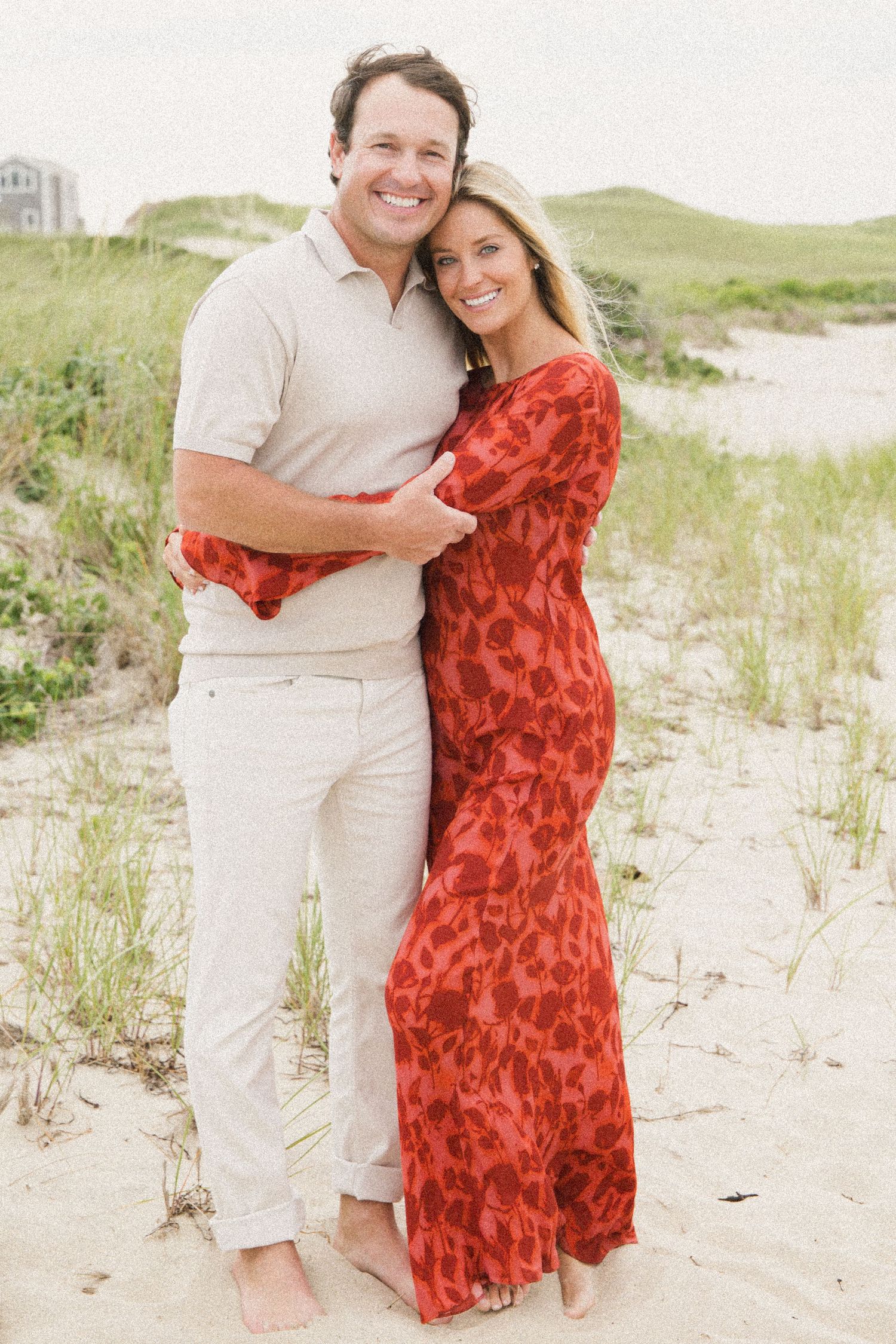A smiling couple embraces on a sandy beach, woman in red floral maxi dress, man in beige polo and white pants.