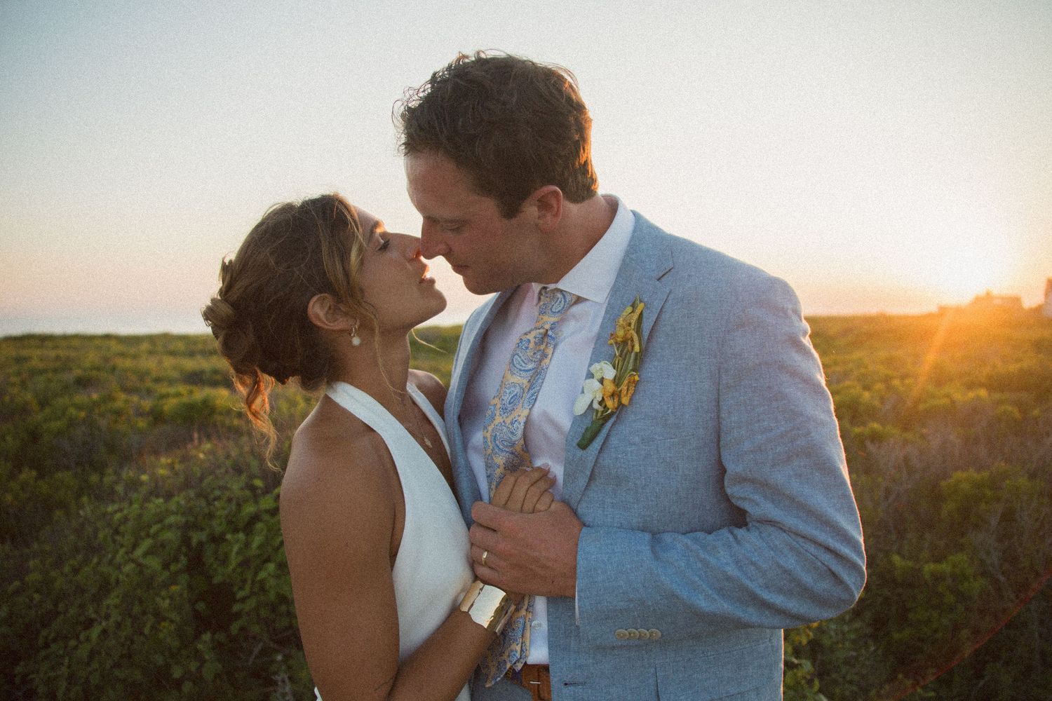 A couple shares an intimate moment against a sunset backdrop on coastal terrain.