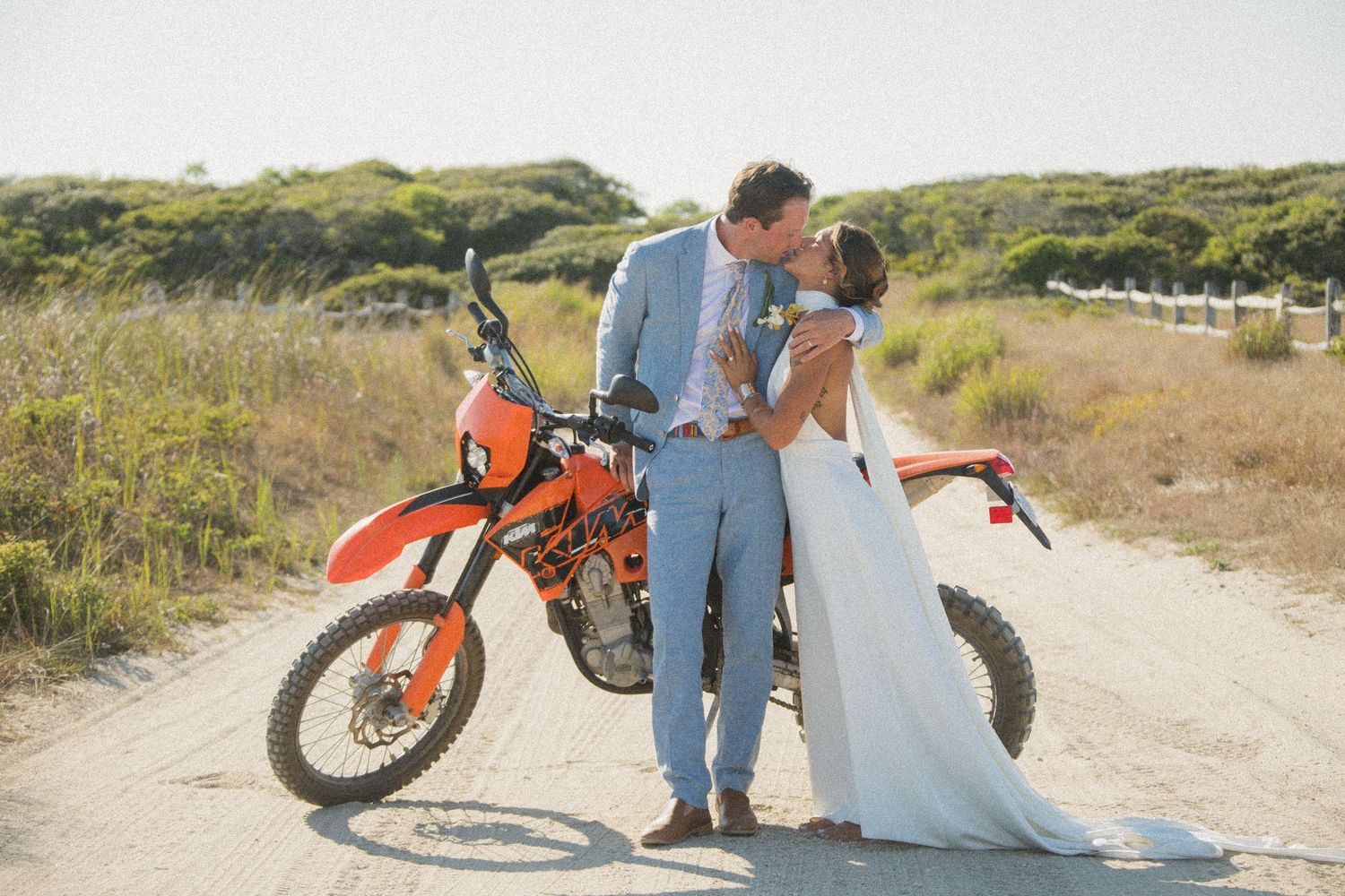 Newlyweds share romantic moment next to orange motorcycle on dirt road surrounded by coastal scenery.