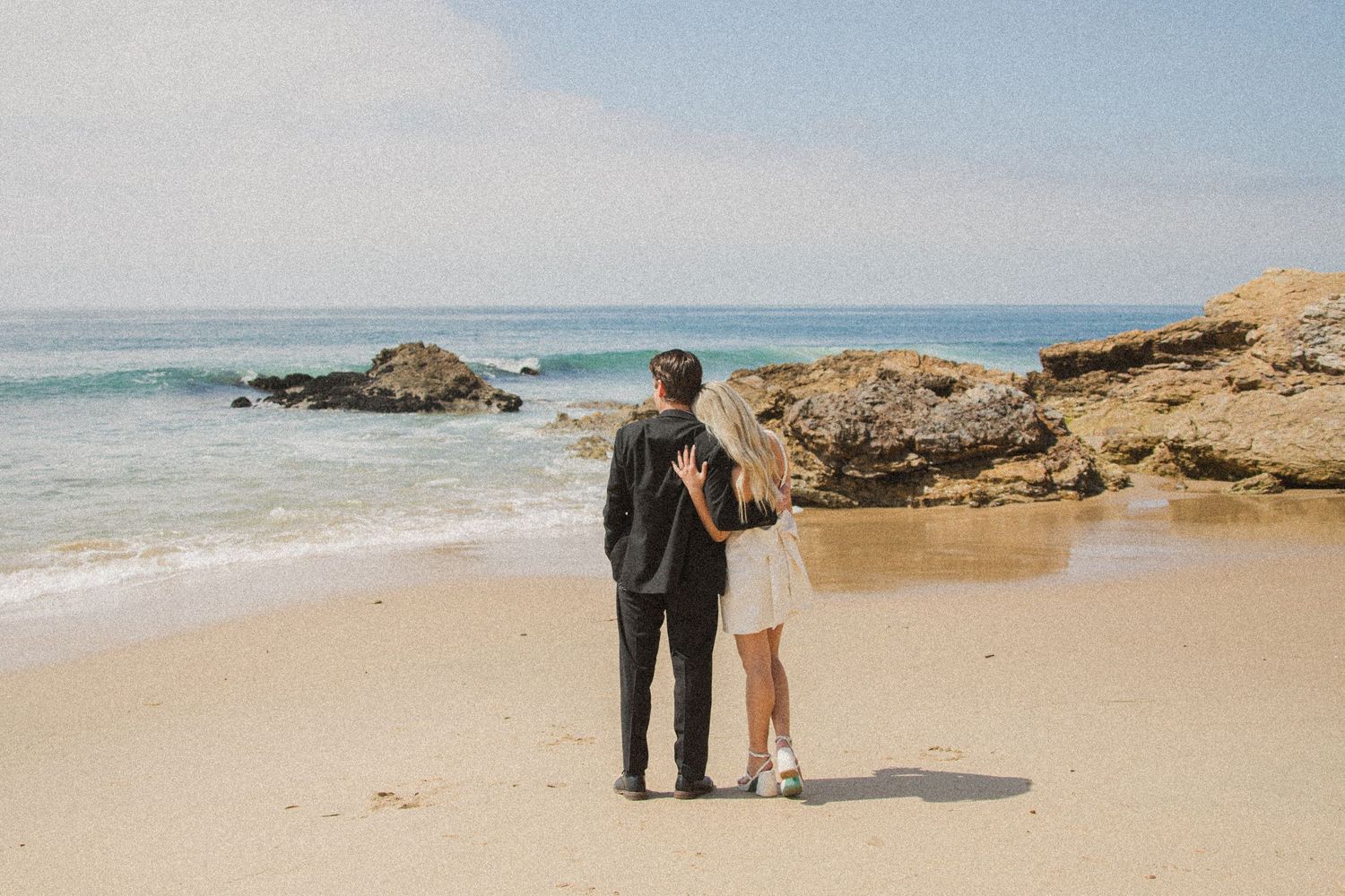 Distant view of wedding couple walking together along scenic rocky beach.