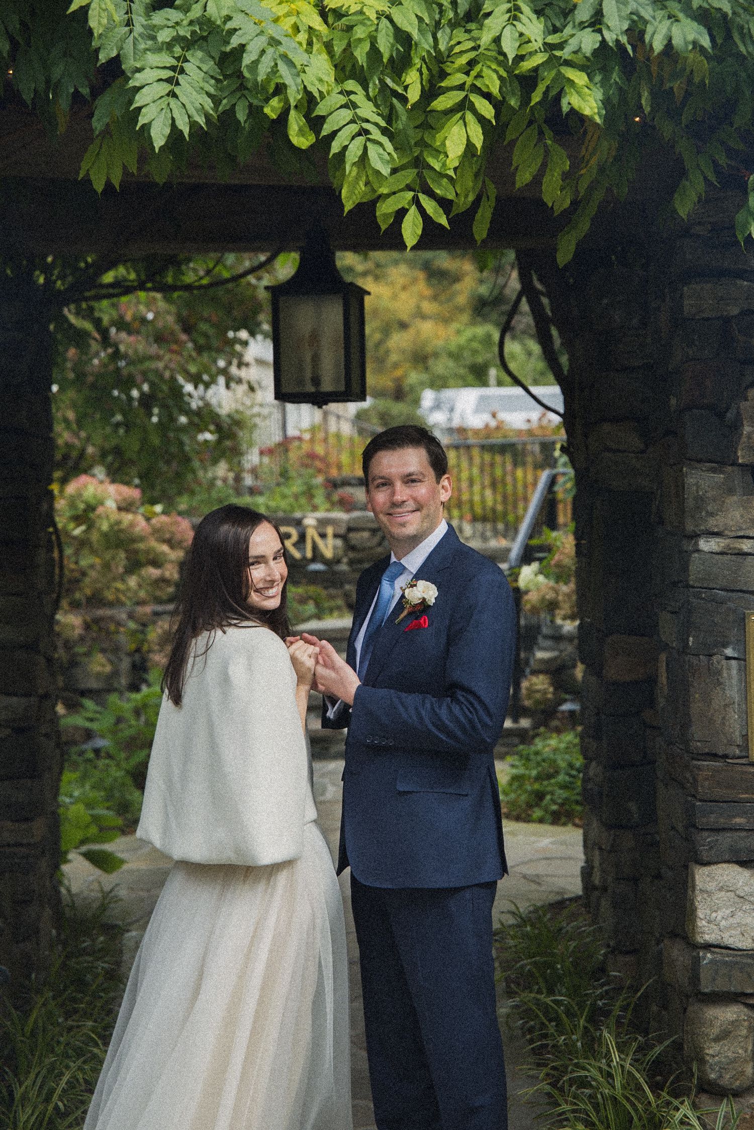 Smiling bride in a white dress and shawl holds hands with a groom in a navy suit beneath a stone archway with hanging lantern and greenery.