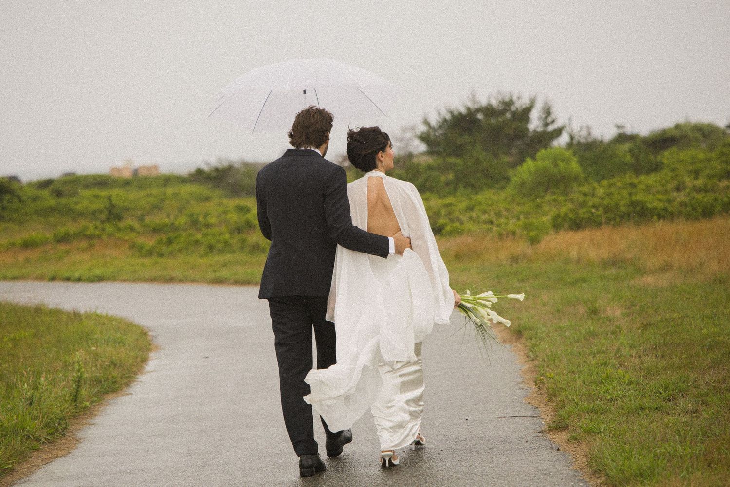 Bride and groom walking hand in hand under a white umbrella on a rainy wedding day outdoors.