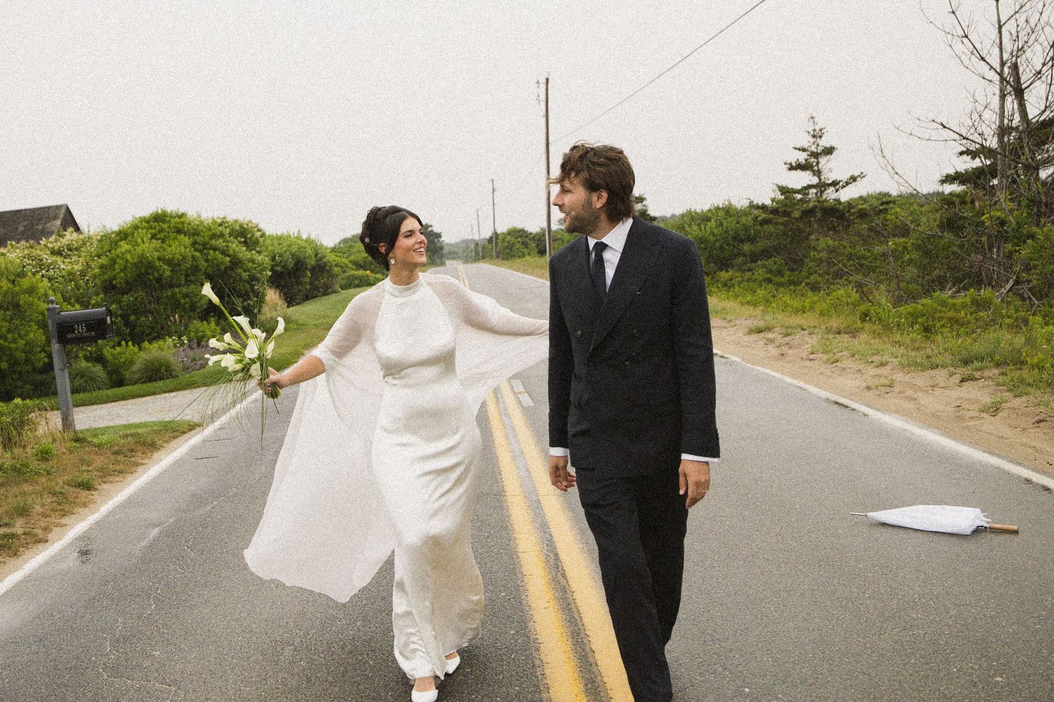Bride and groom walk hand-in-hand down a quiet road on their wedding day, bride in white gown with veil flowing.