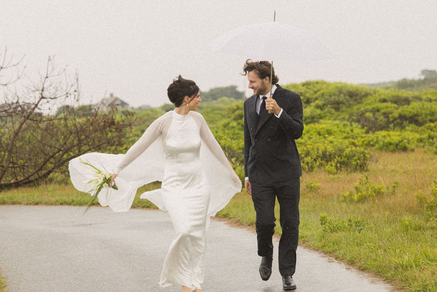 Bride and groom walking on a path in the rain, bride in white gown holding bouquet, groom holding white umbrella.