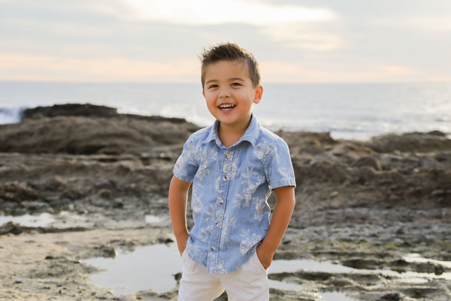 Young boy in a light blue shirt and white pants smiles while standing on rocky beach terrain.