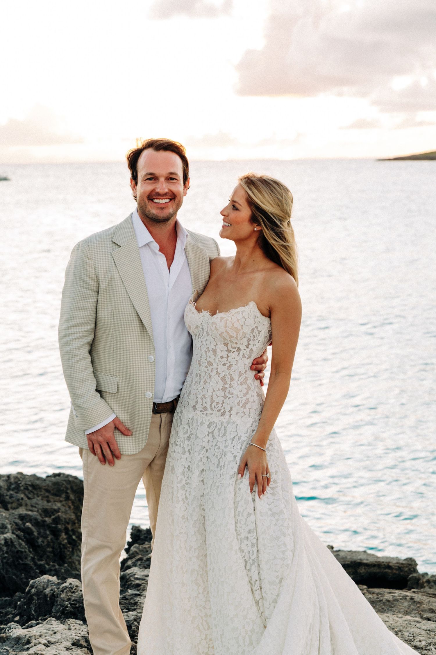 Couple in wedding attire poses against ocean backdrop on rocky coastline at sunset.