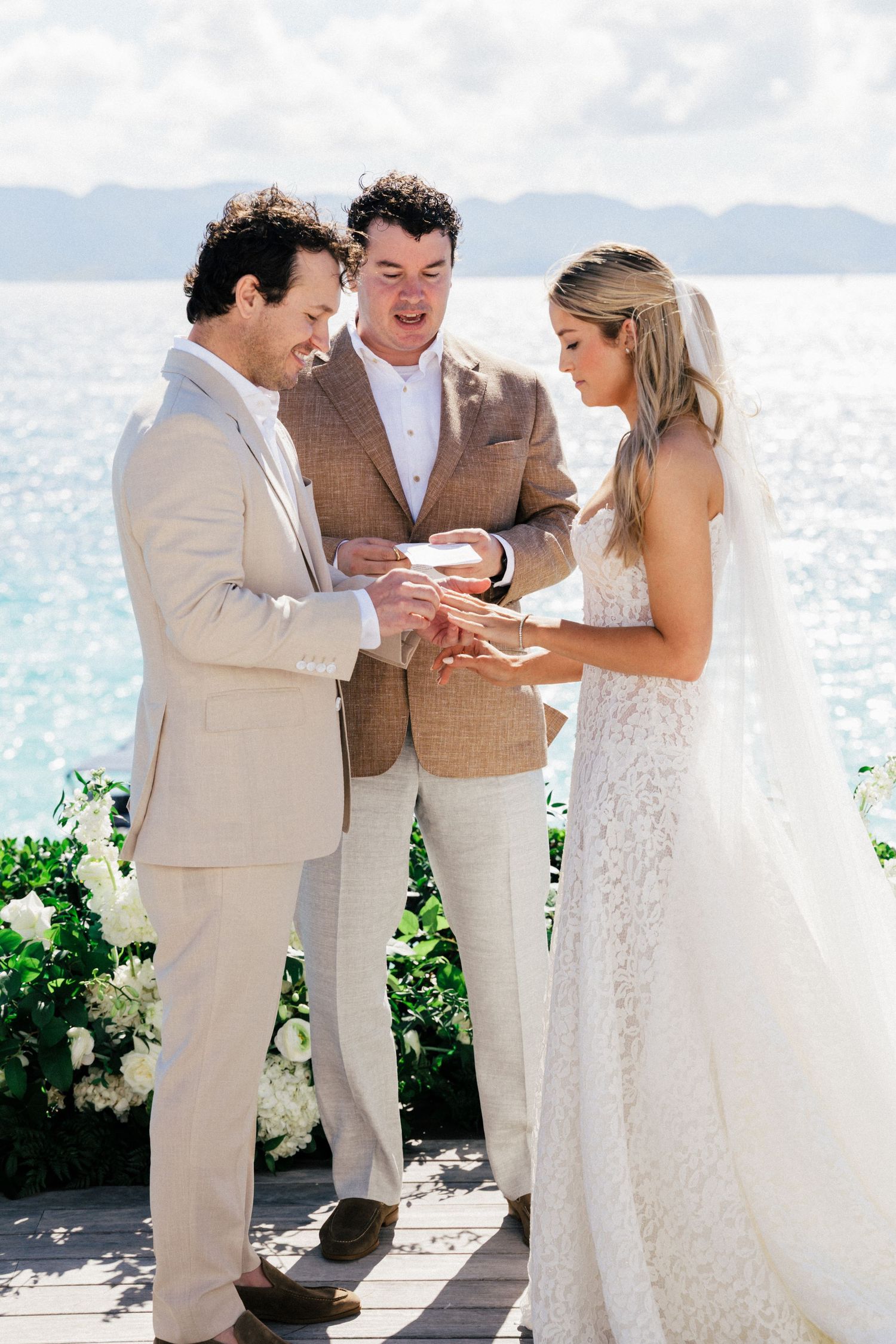 Wedding ceremony participants share a touching moment on a seaside terrace with white flowers.