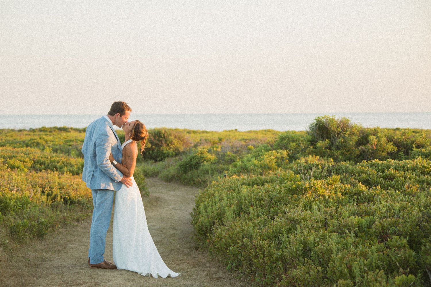 A couple in formal attire embraces on a scenic coastal path surrounded by wildflowers at sunset.