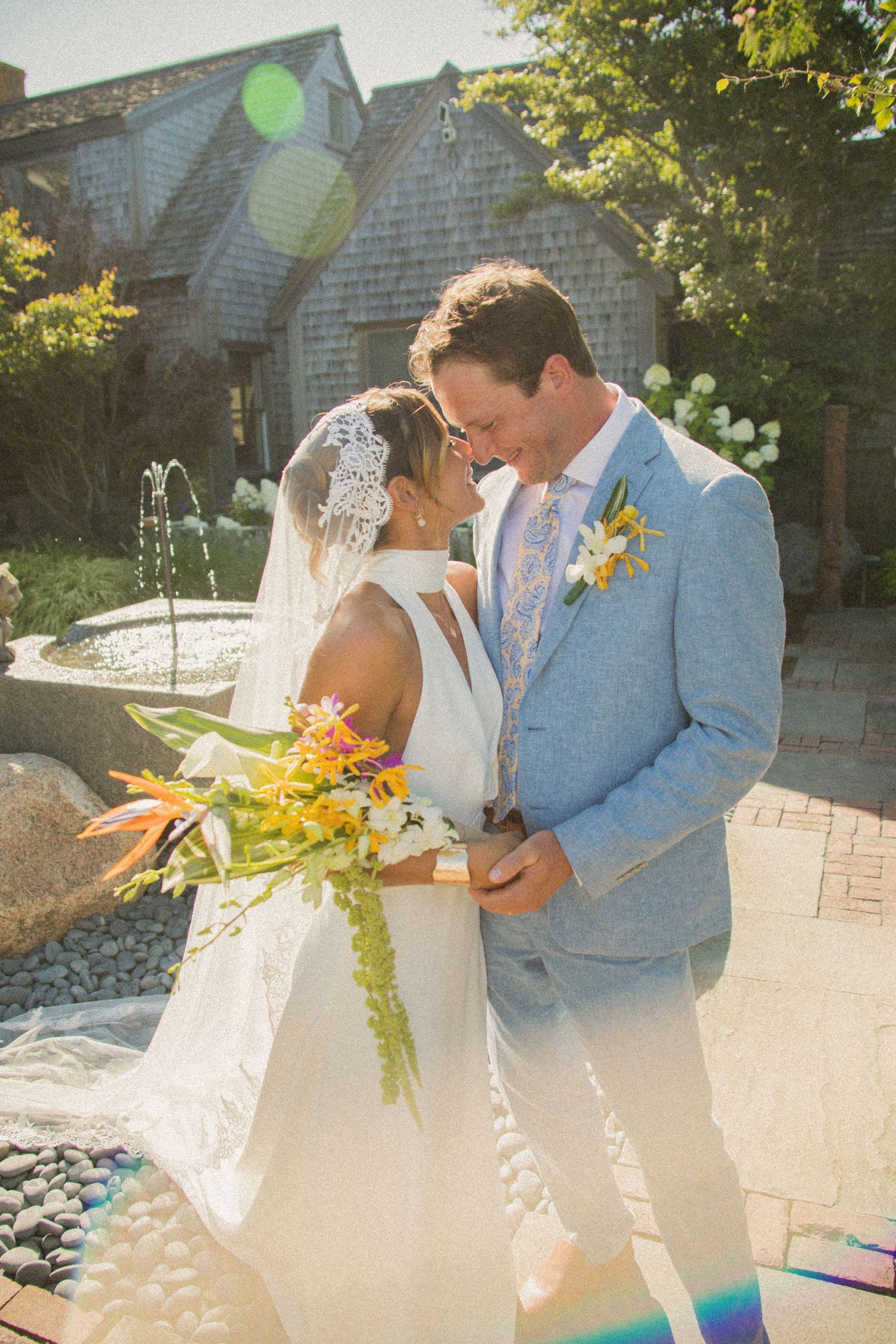 Newlywed couple shares intimate moments in a sunlit garden setting with beautiful floral bouquet and light lens flares.