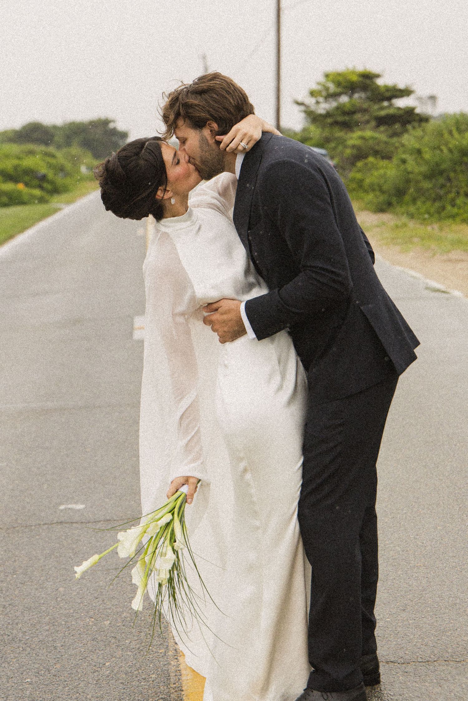A bride and groom share a romantic kiss on a road, she holds white calla lilies in a flowing white dress.