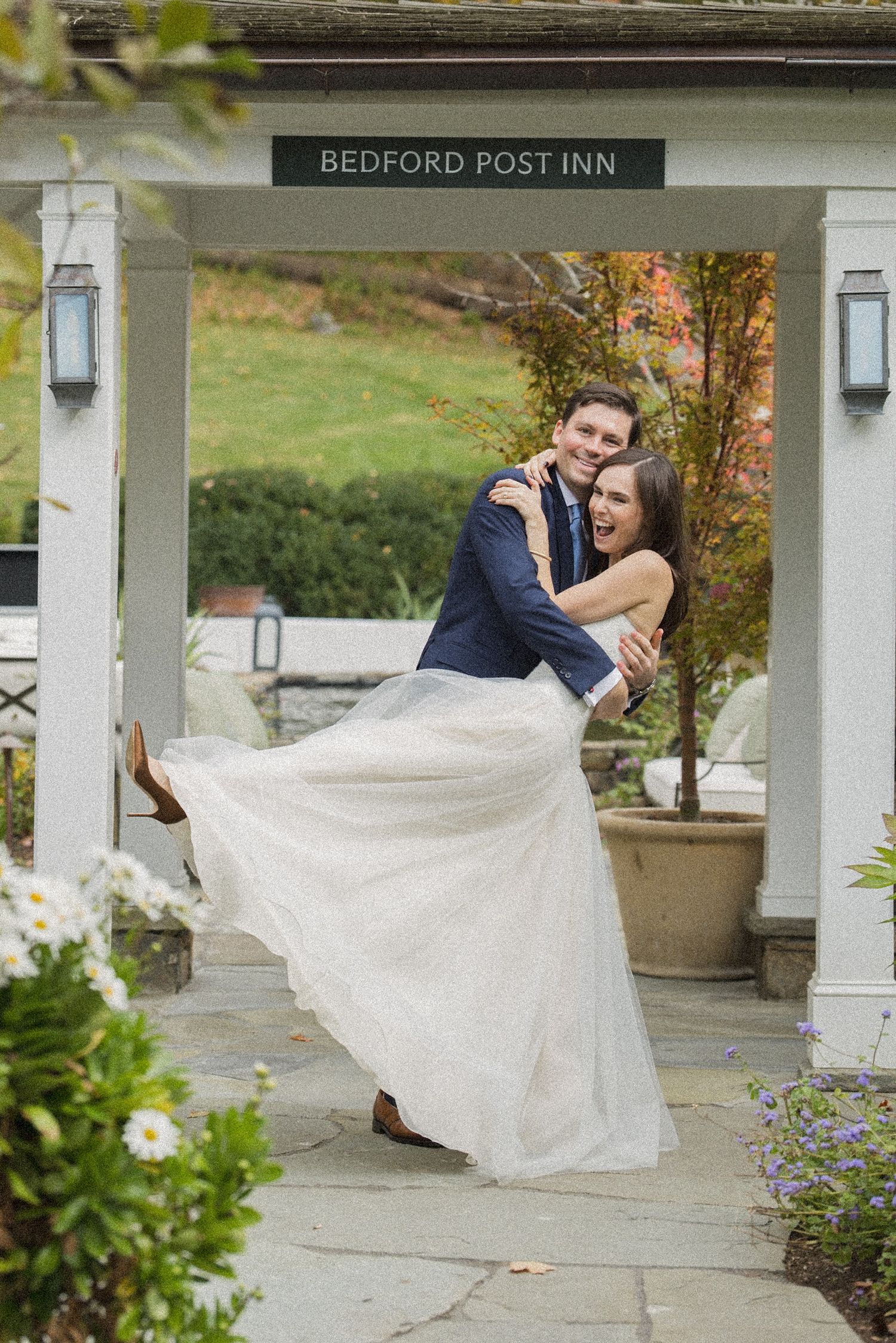 Groom lifts his laughing bride in a tulle wedding gown under the Bedford Post Inn entrance, surrounded by autumn garden greenery.