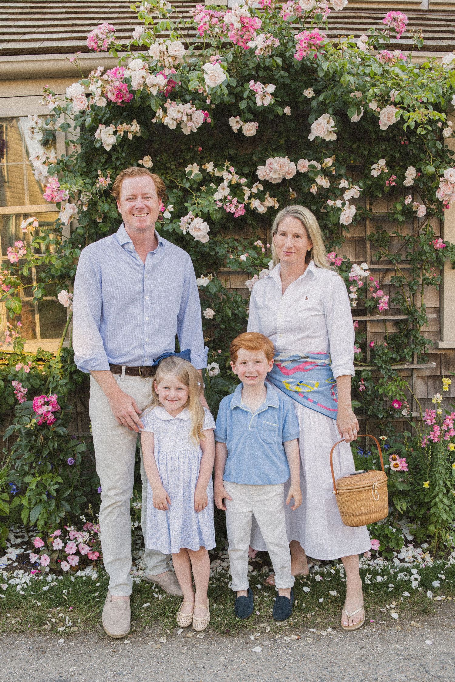 Family of four posing in front of a floral arch with pink roses in a garden setting.