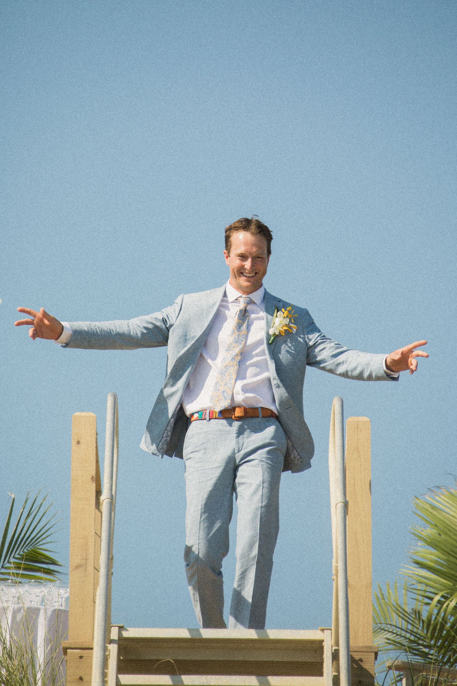 Groomsman in light blue suit performs playful jumping poses on wooden boardwalk at beach wedding.
