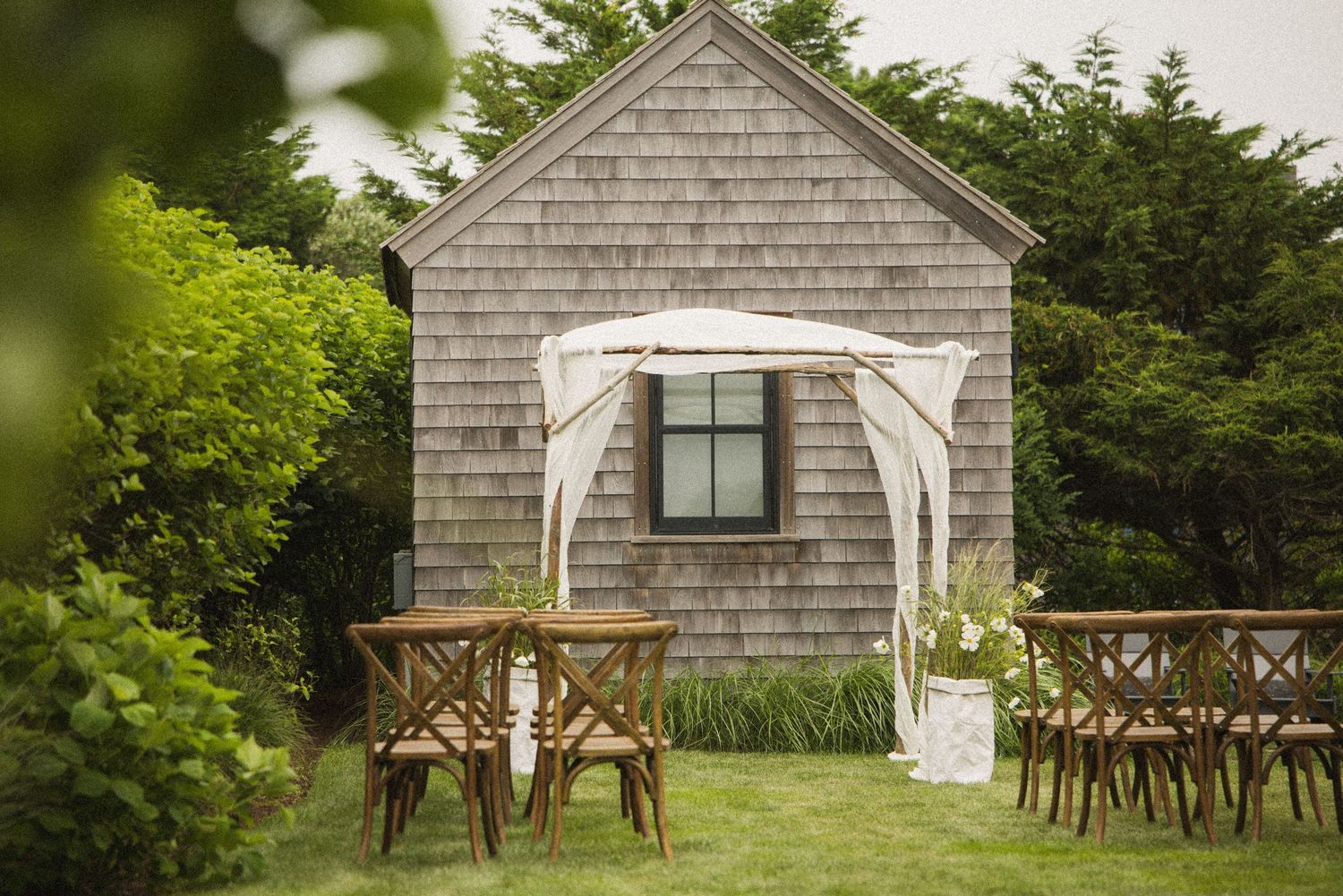 Outdoor wedding ceremony setup with wooden chairs and white draped arch on green lawn beside rustic gray shingled cottage.
