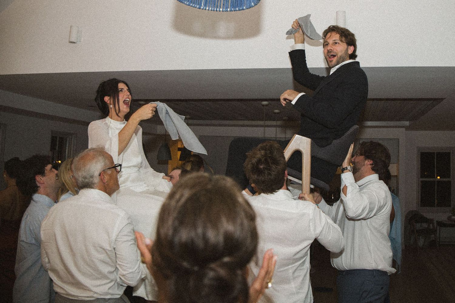 Bride and groom lifted on chairs during traditional Hora dance at lively wedding reception celebration.