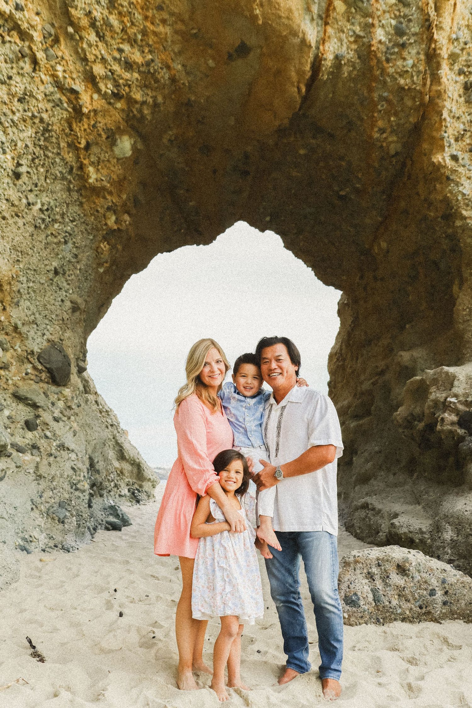 A family poses together under a natural rock arch formation at the beach during golden hour.