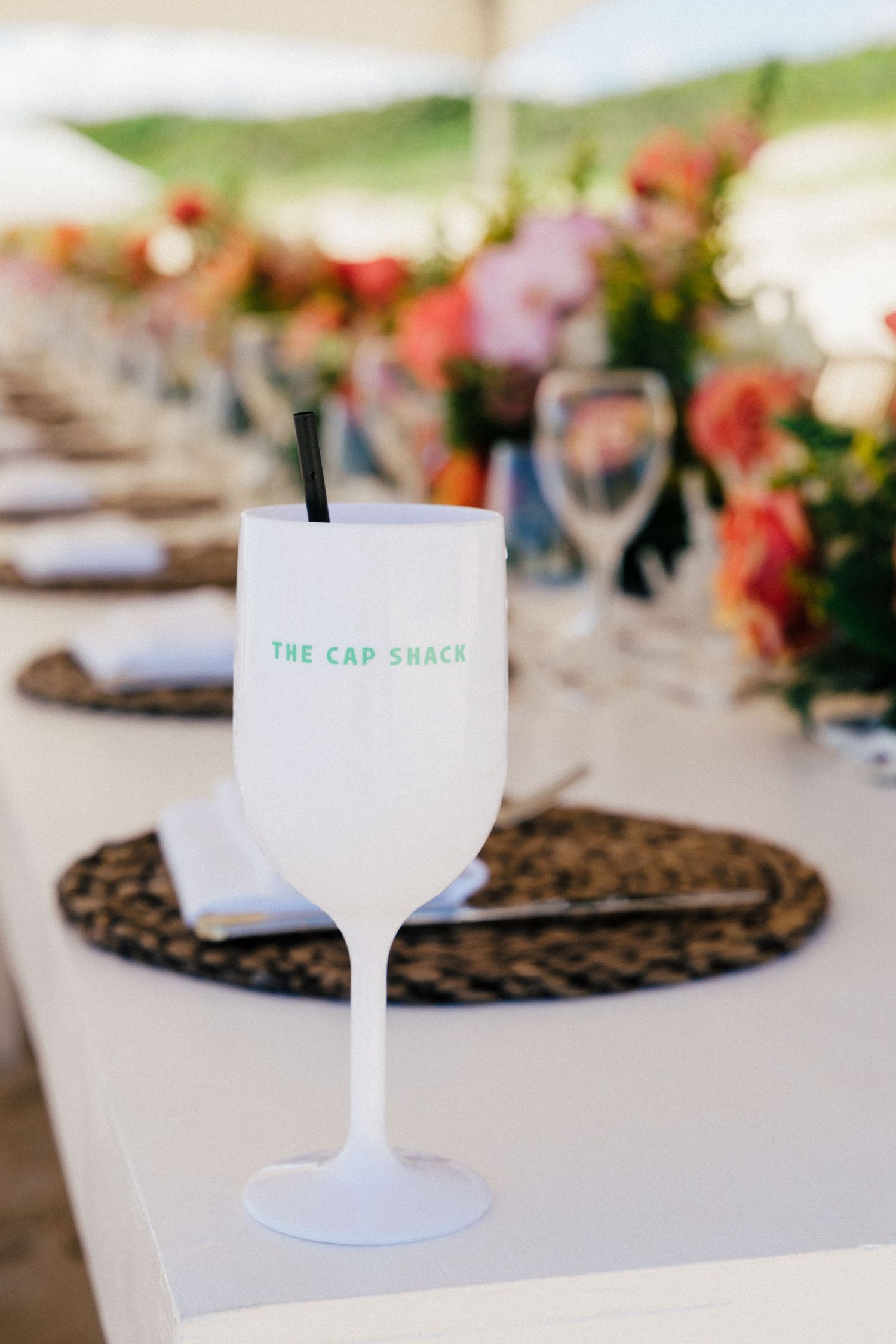 A frosted white wine glass with 'The Cap Shack' branding sits on a woven placemat at an elegant table setting.