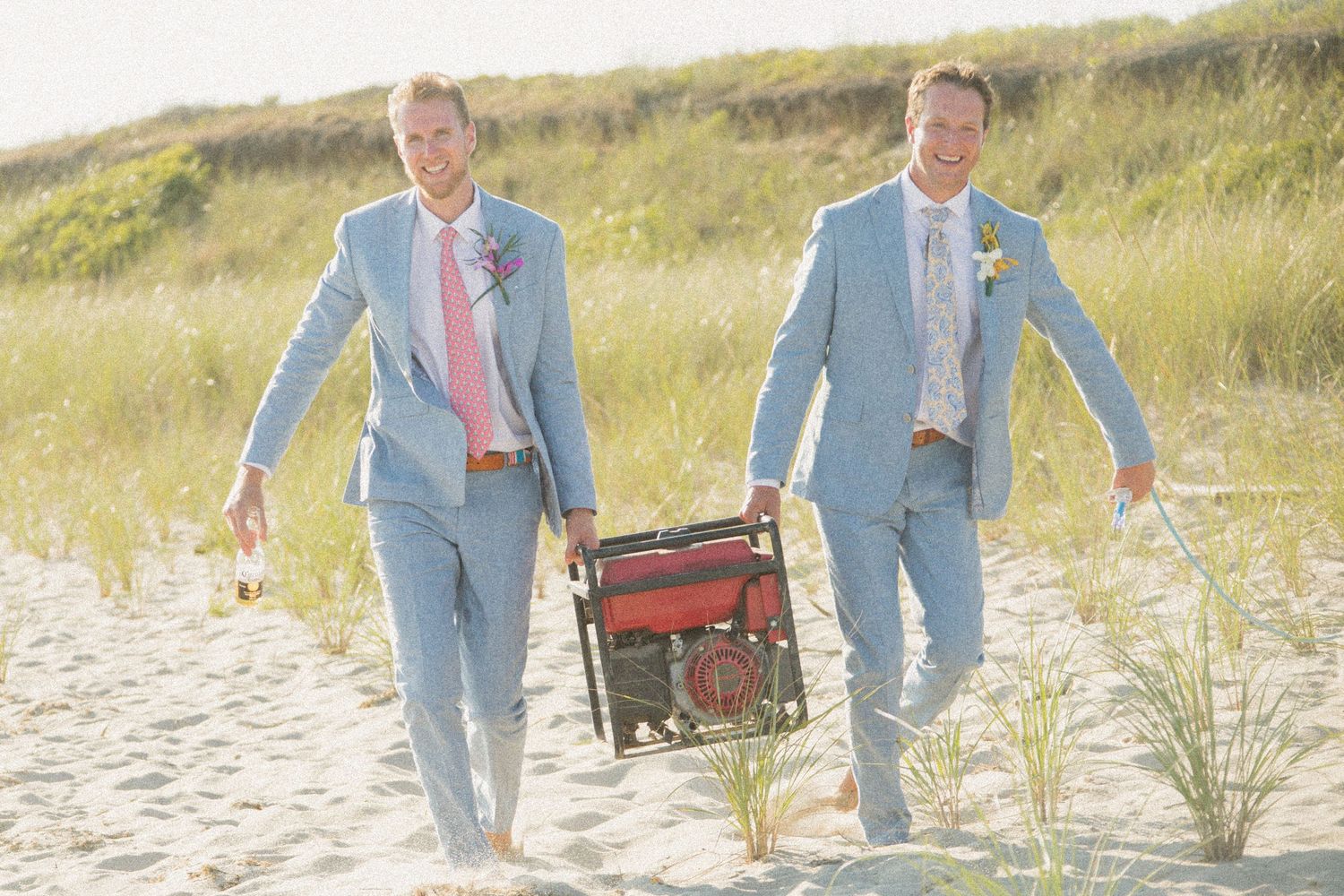 Two people in matching light blue suits carrying vintage luggage across beach dunes.