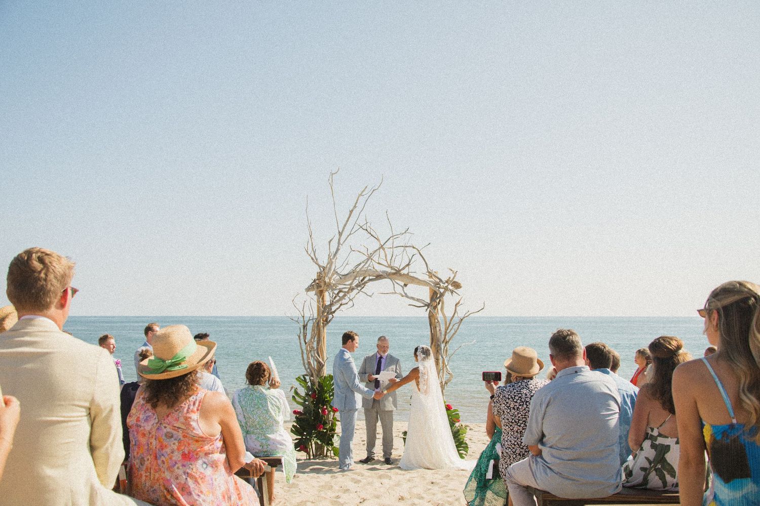 Beach wedding ceremony with guests watching from their seats by the ocean.