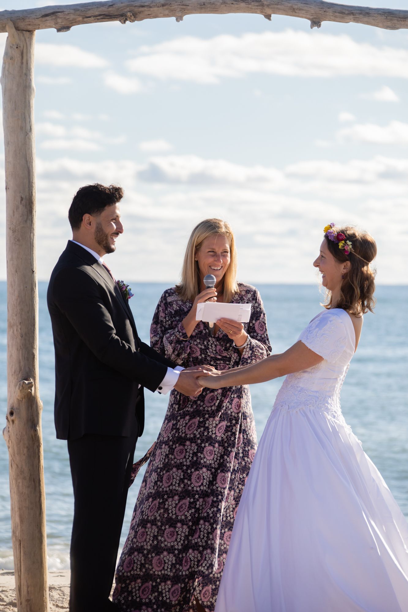 A wedding ceremony under a rustic wooden arch on a sunny beach.