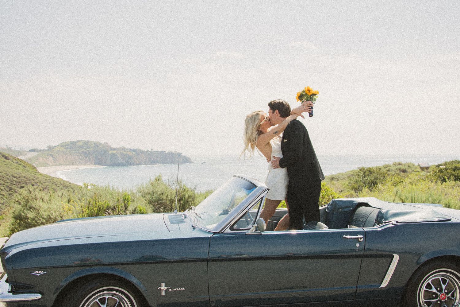 Romantic couple riding in vintage convertible car along coastal road at sunset.
